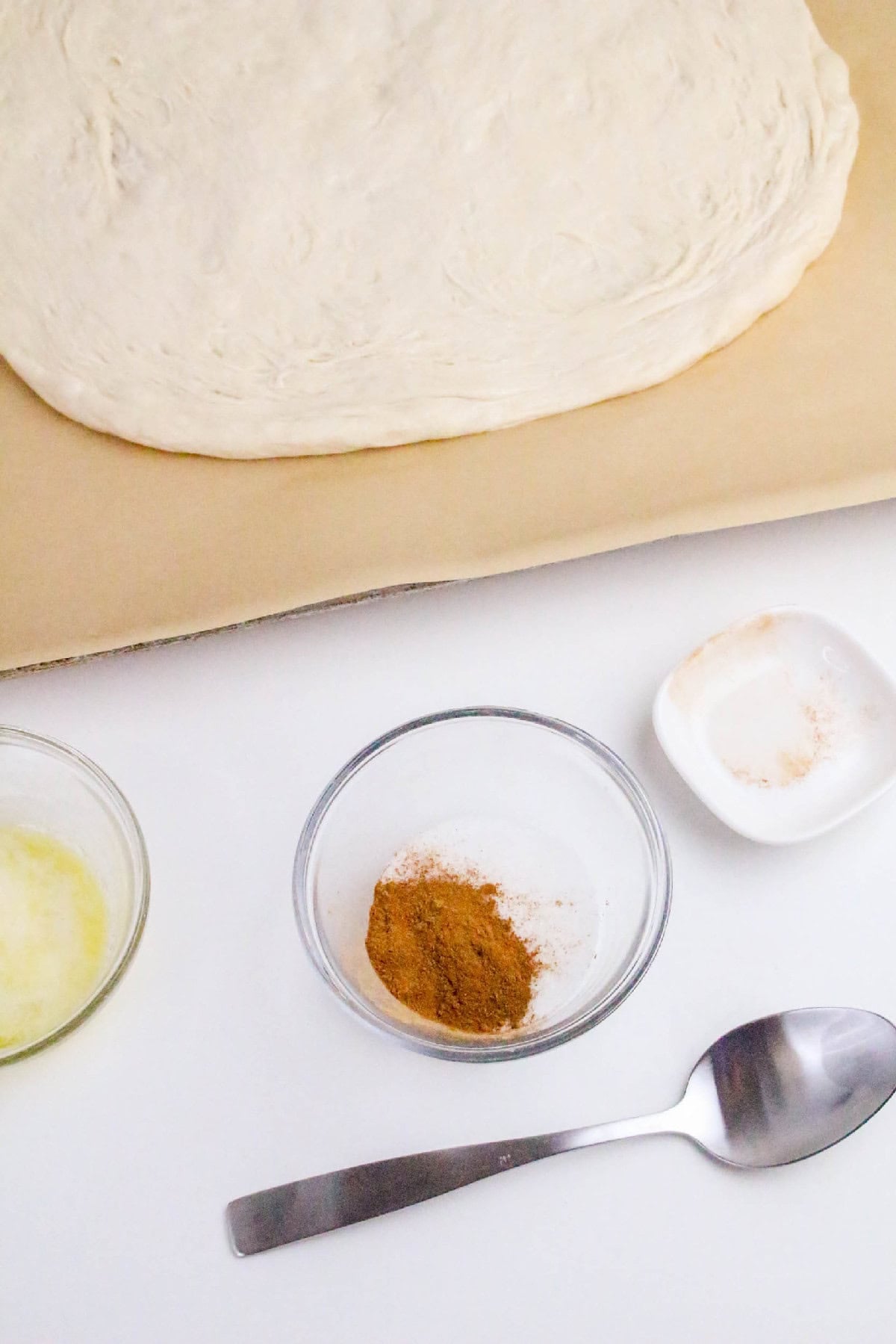 Close-up of pizza dough on parchment paper with cinnamon sugar, melted butter, and spoon prepared for Nutella pizza topping.