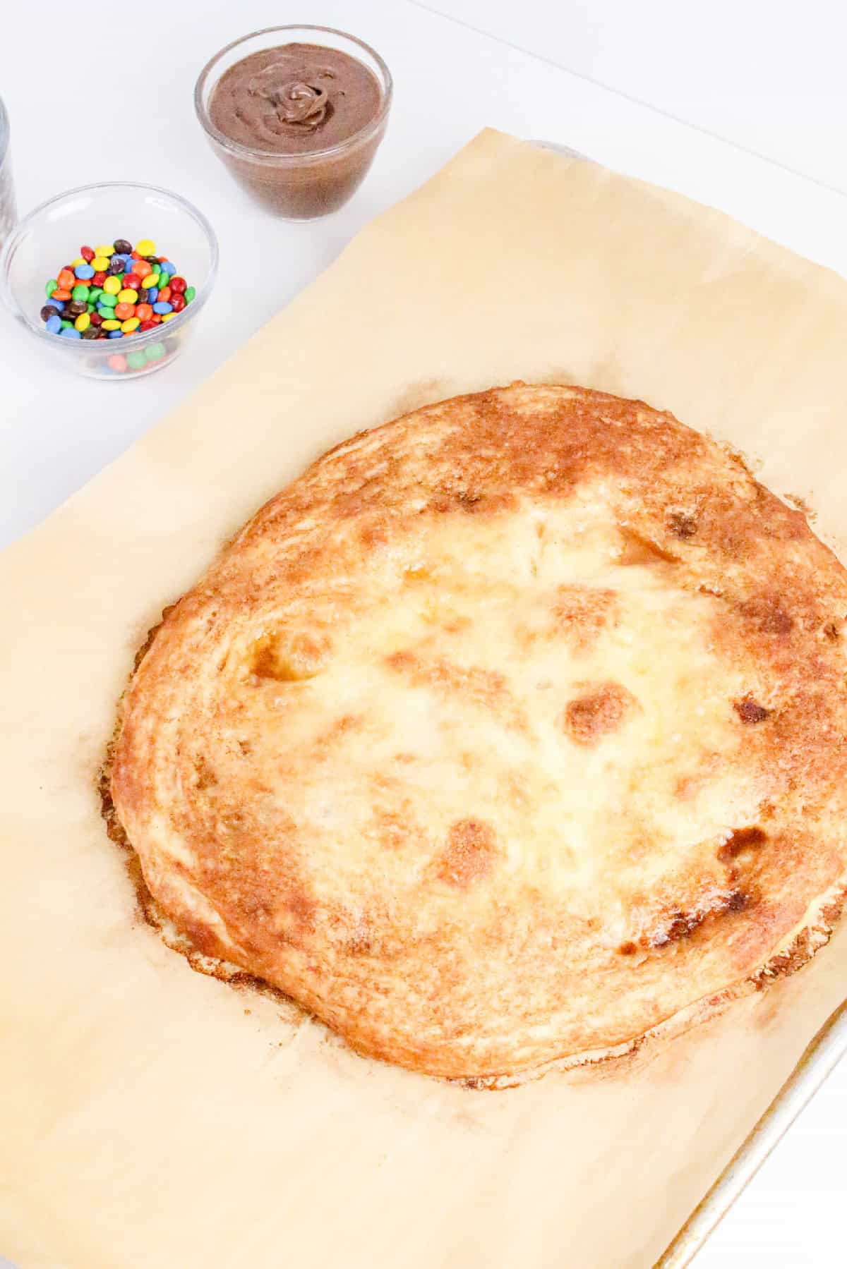 Freshly baked pizza crust on parchment paper with Nutella and candy toppings prepared nearby for a dessert pizza.
