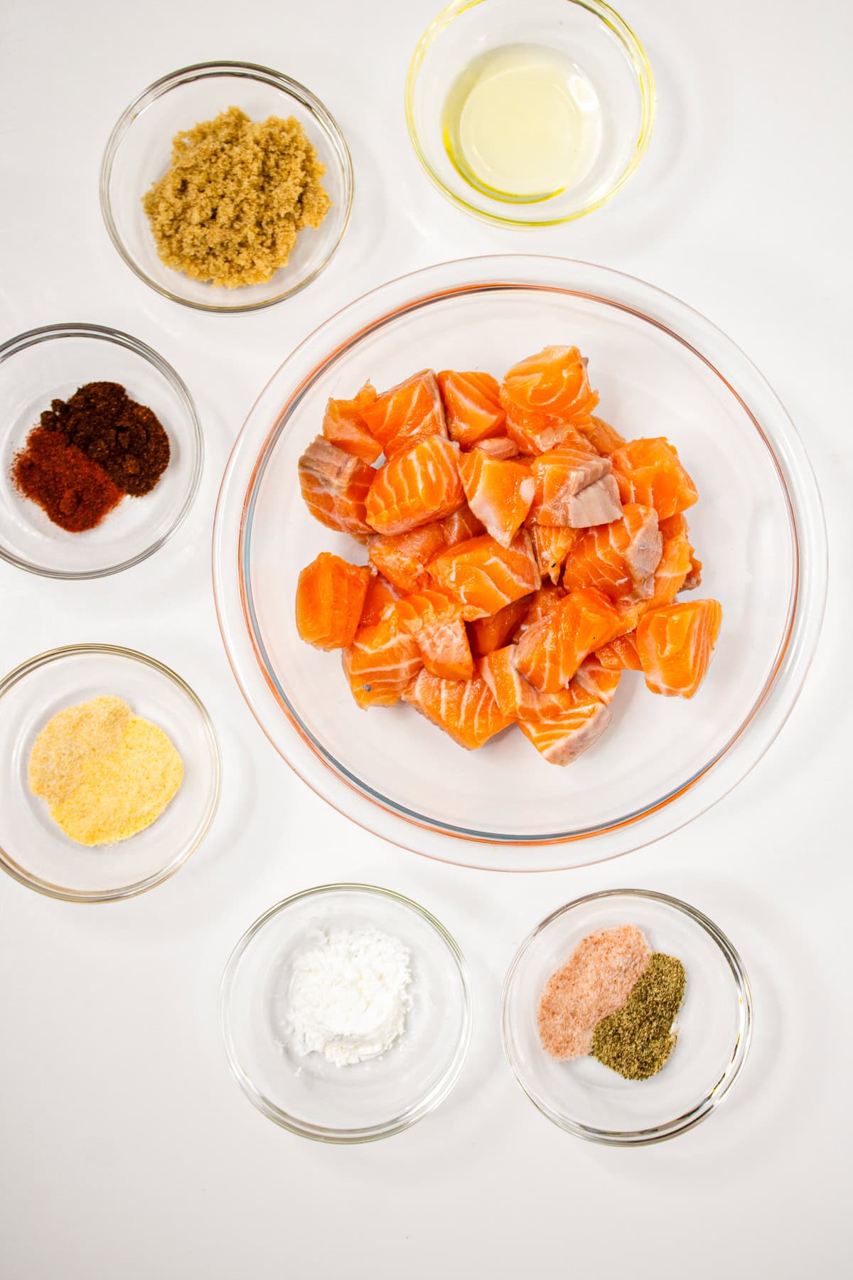 Air fryer salmon bites ingredients including cubed salmon, brown sugar, paprika, garlic powder, cornstarch, salt, pepper, and oil in small bowls on a white background.