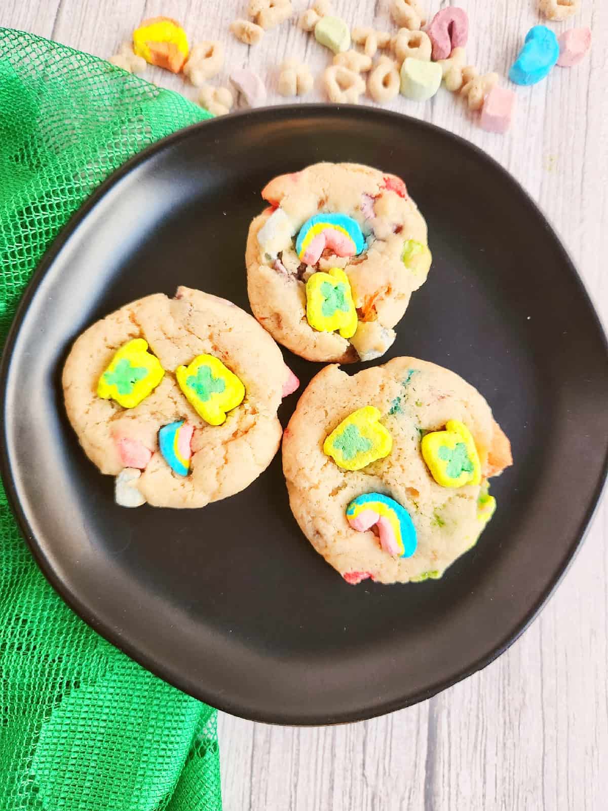 Soft Lucky Charms cookies topped with rainbow and shamrock marshmallows on a black plate.