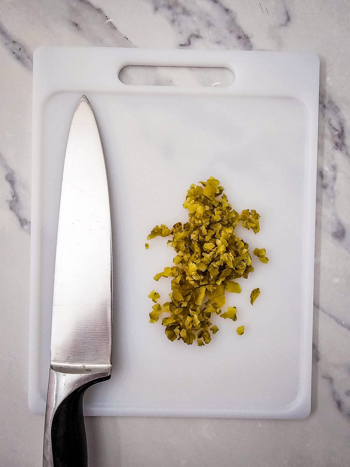 Finely chopped dill pickles on a white cutting board with a chef’s knife.