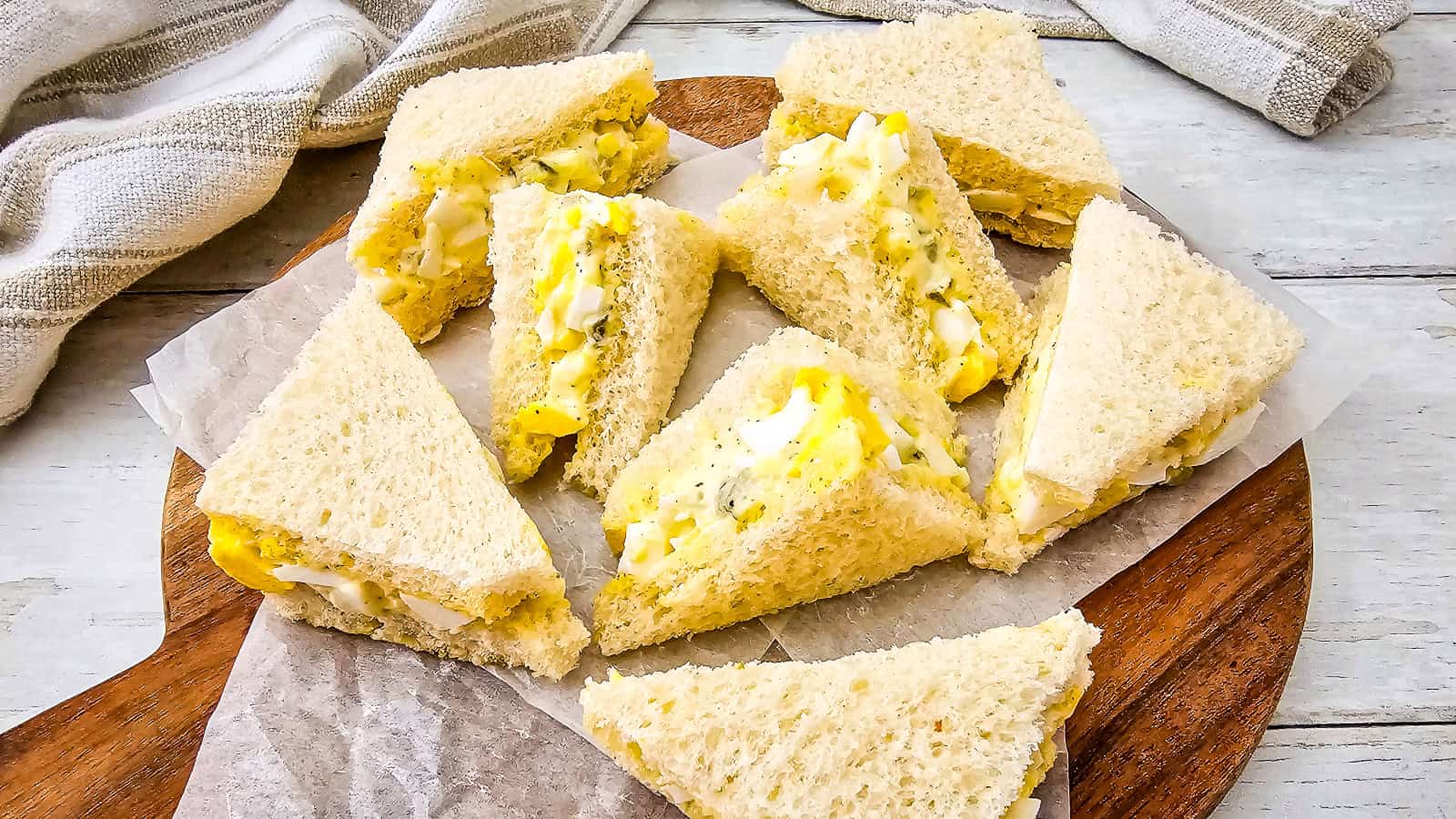 Egg salad tea sandwiches arranged on a wooden board with parchment paper, served with soft white bread and creamy filling.