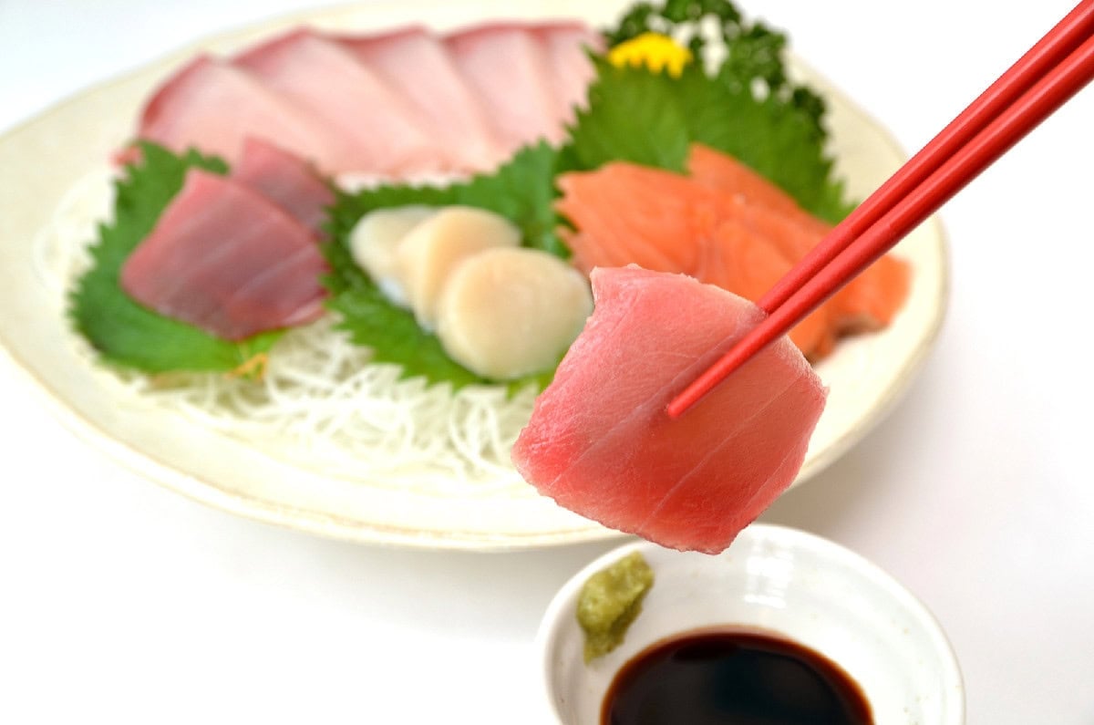 Fresh tuna sashimi held with chopsticks over a plate with assorted sashimi, soy sauce, and wasabi.