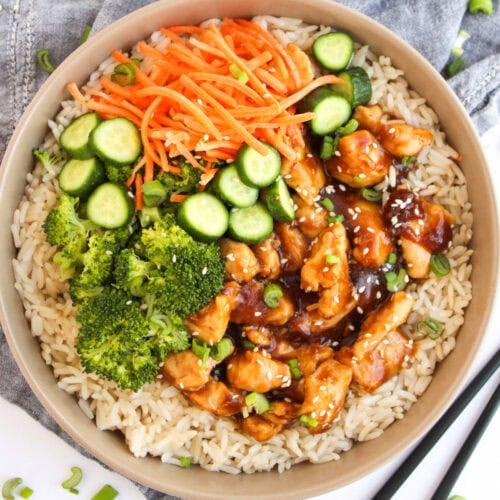 Teriyaki chicken rice bowl with broccoli, cucumber slices, shredded carrots, and sesame seeds on a white background.