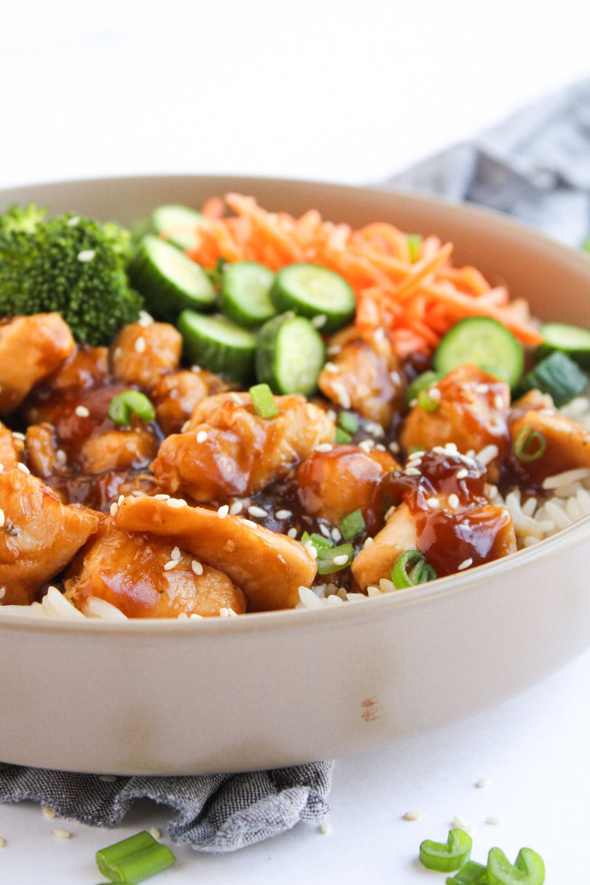 Close-up of teriyaki chicken over rice with cucumbers and carrots in a bowl.