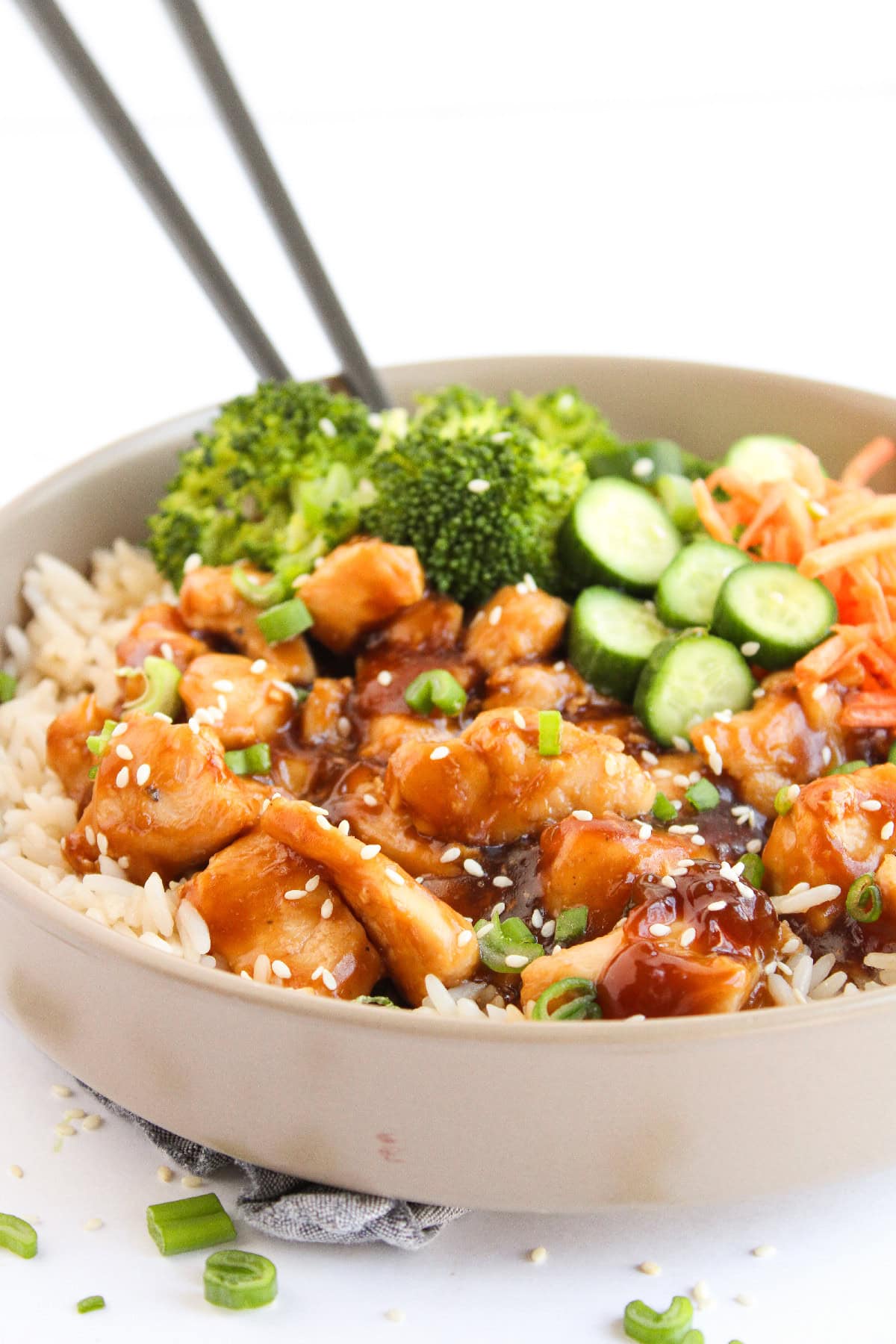 Close-up of teriyaki chicken over rice with cucumbers and carrots in a bowl.
