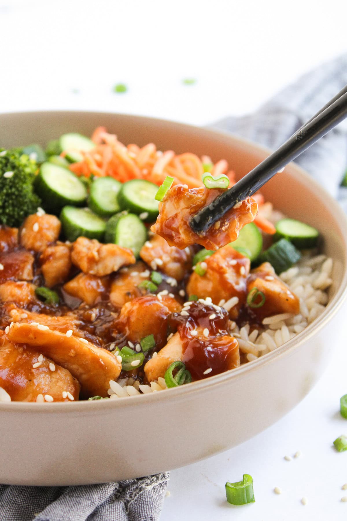 Chopsticks holding teriyaki chicken over a rice bowl with broccoli and cucumbers.