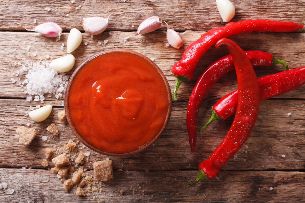 Bowl of sriracha sauce with fresh red chilies, garlic, and brown sugar on a wooden table.