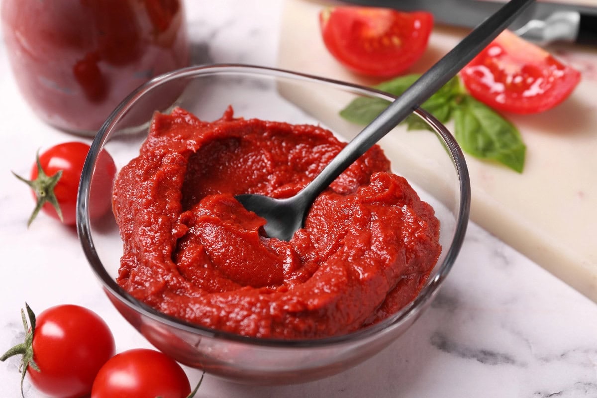 Tomato paste in a glass bowl with spoon and fresh tomatoes nearby.