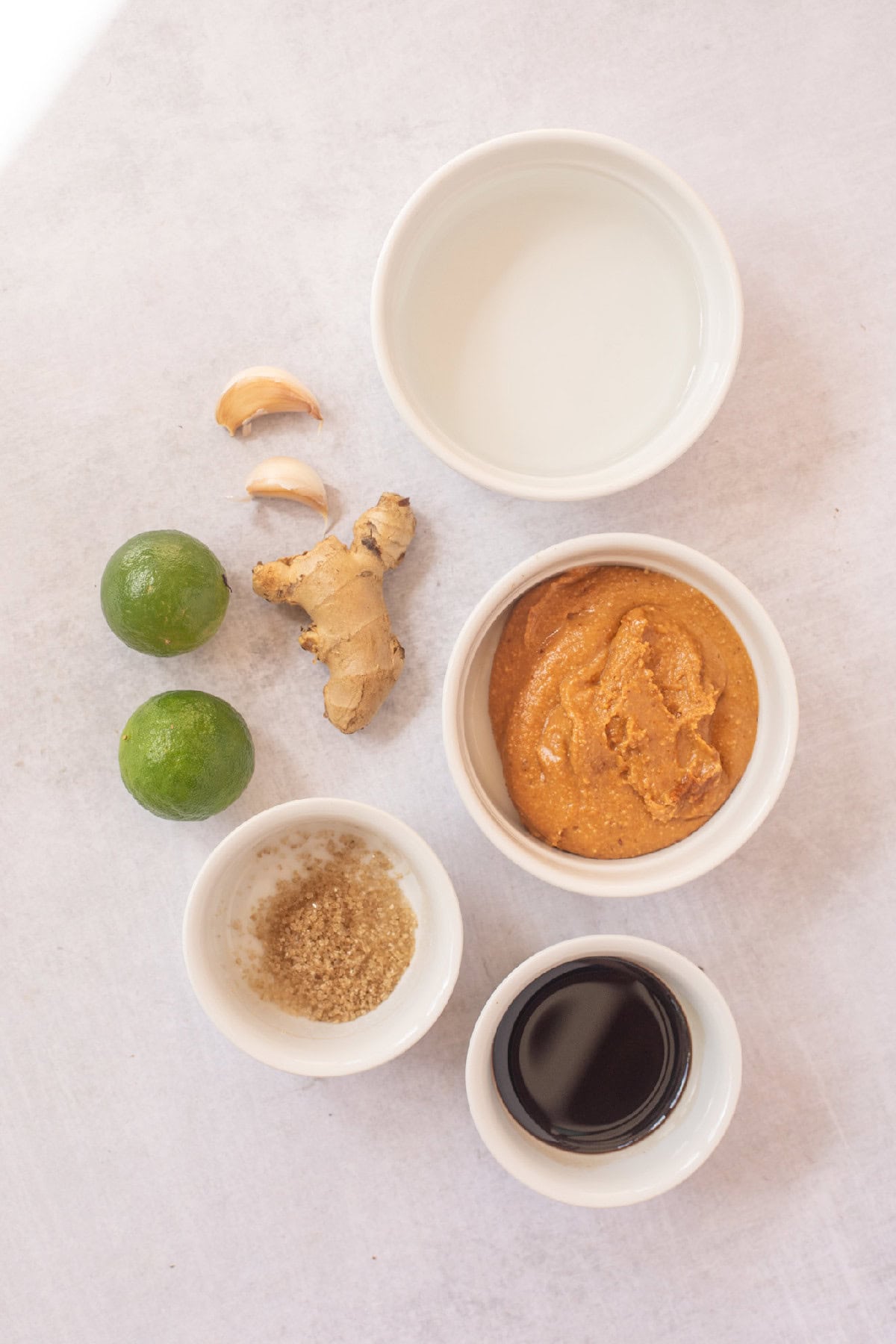 Ingredients for peanut sauce including peanut butter, soy sauce, lime, garlic, ginger, brown sugar, and water in small white bowls.