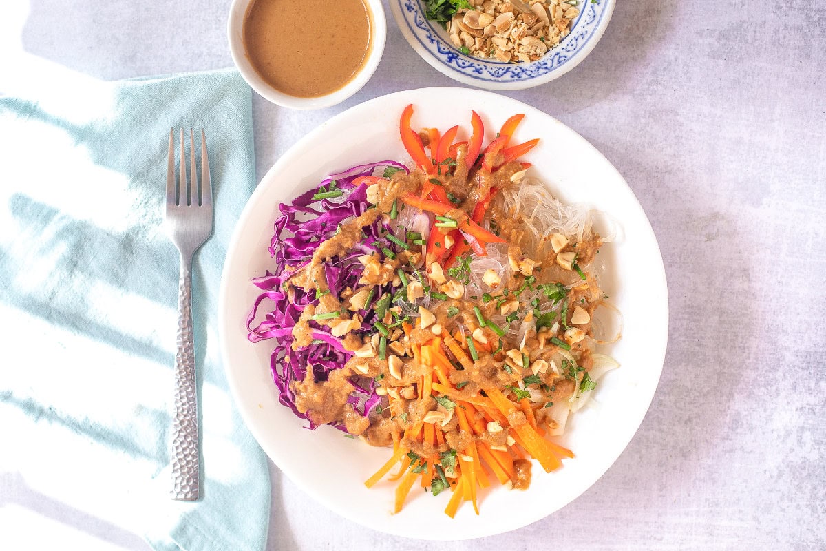 Overhead view of a colorful spring roll in a bowl with peanut sauce, rice noodles, carrots, cabbage, and peanuts.