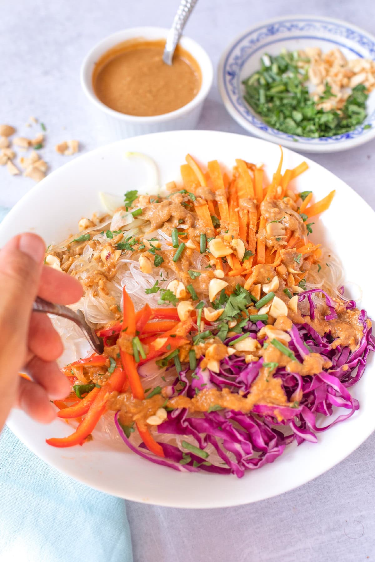 Hand holding a fork over a spring roll bowl with rice noodles, vegetables, chopped peanuts, and peanut sauce.
