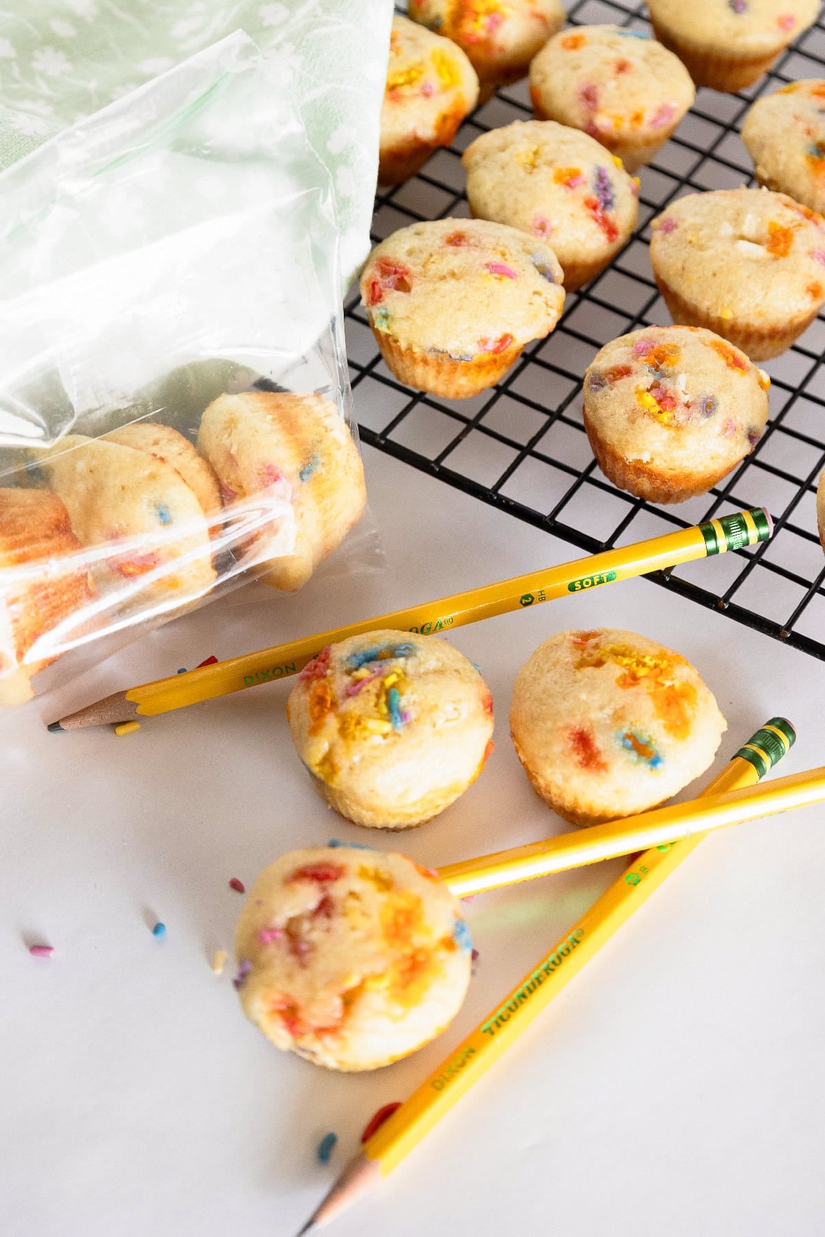 Birthday cake mini muffins beside yellow pencils and a plastic storage bag on a white surface.