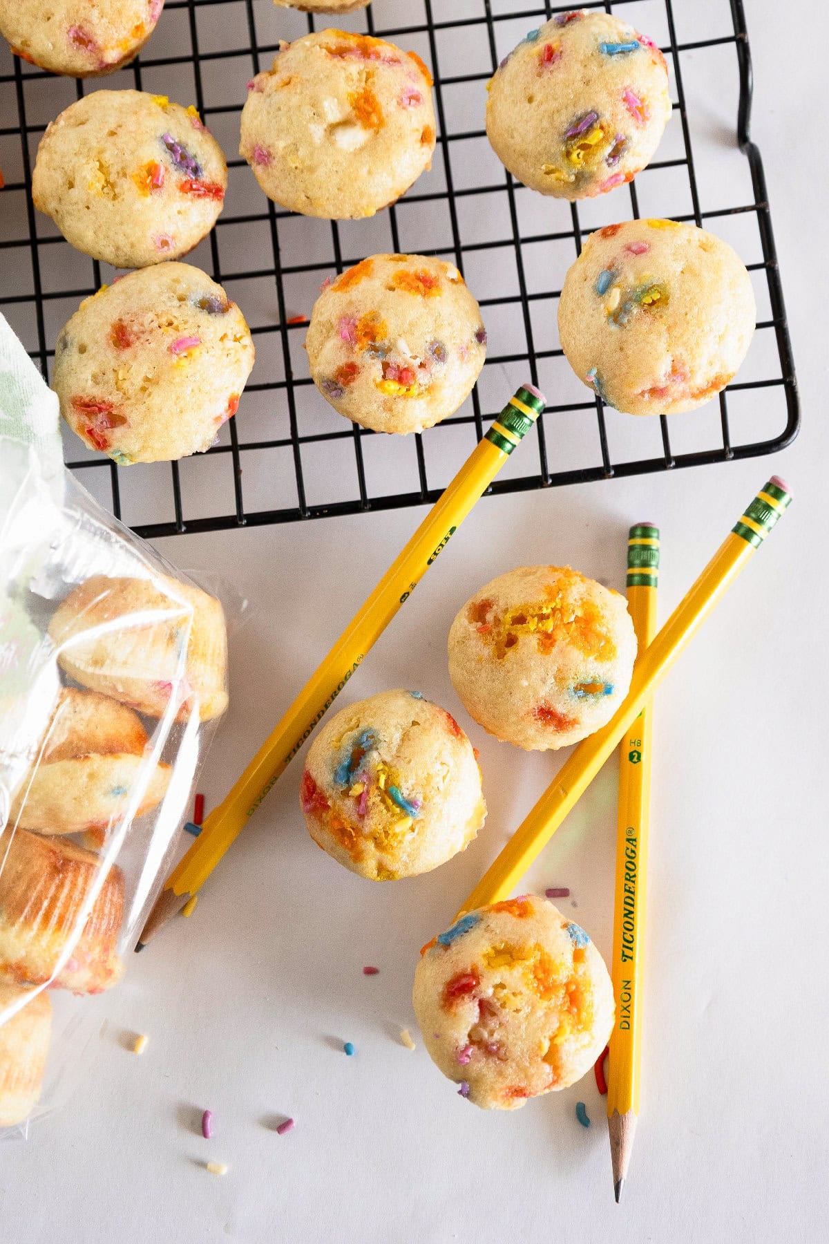 Overhead view of birthday cake mini muffins with pencils, rainbow sprinkles, and a cooling rack.