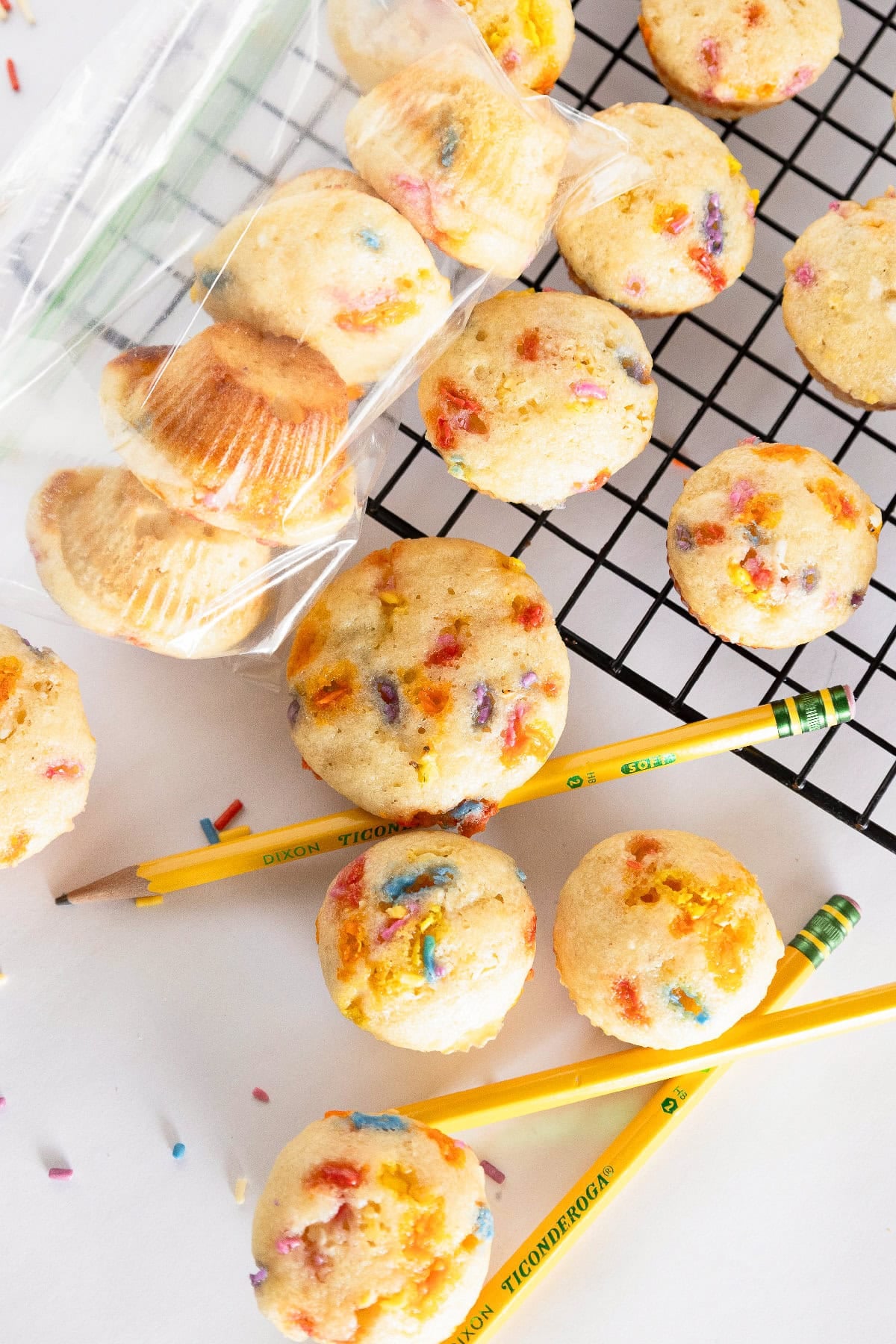 Birthday cake mini muffins with colorful sprinkles arranged around yellow pencils and a cooling rack.