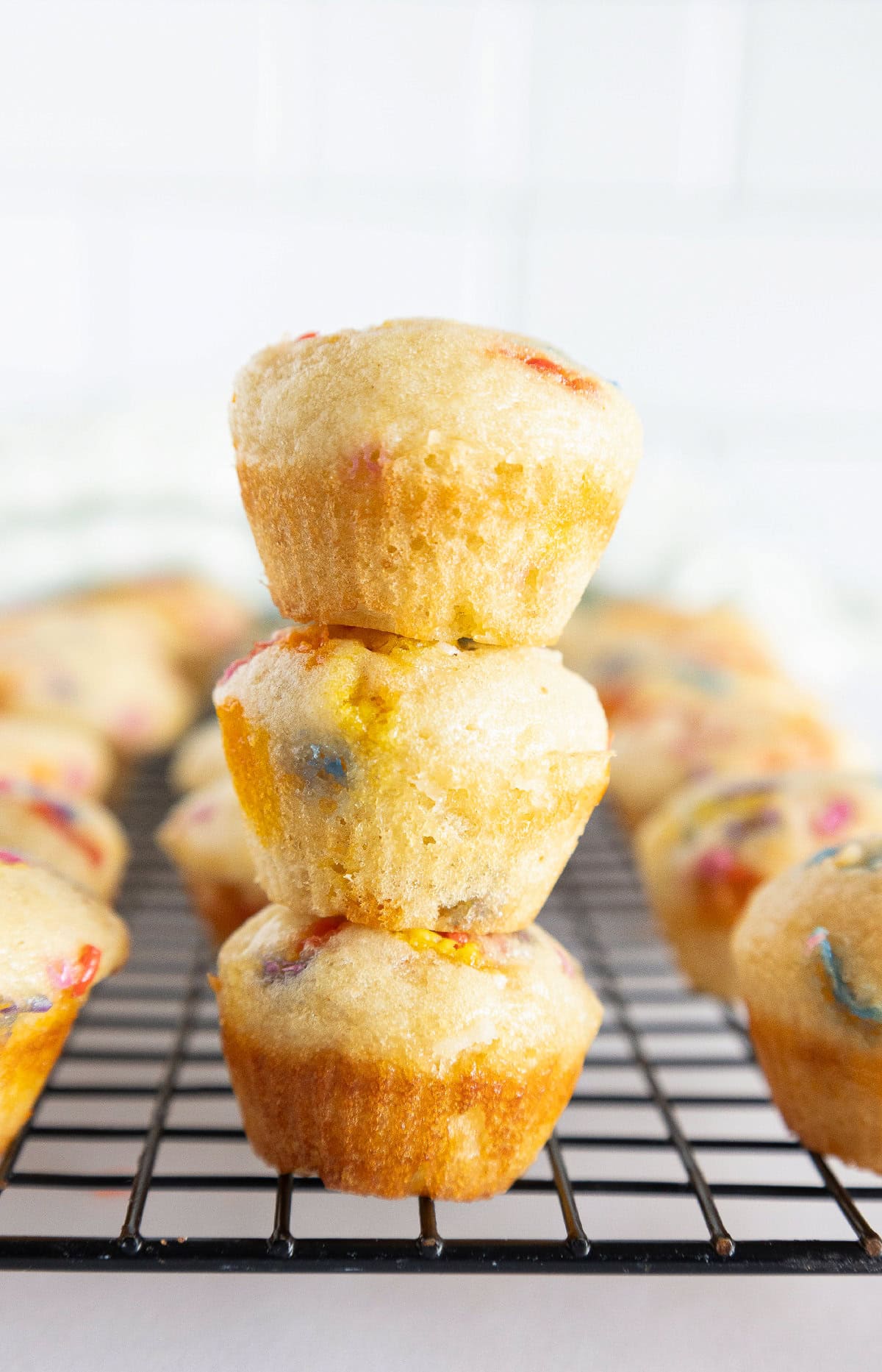 Three stacked birthday cake mini muffins with rainbow sprinkles on a cooling rack.