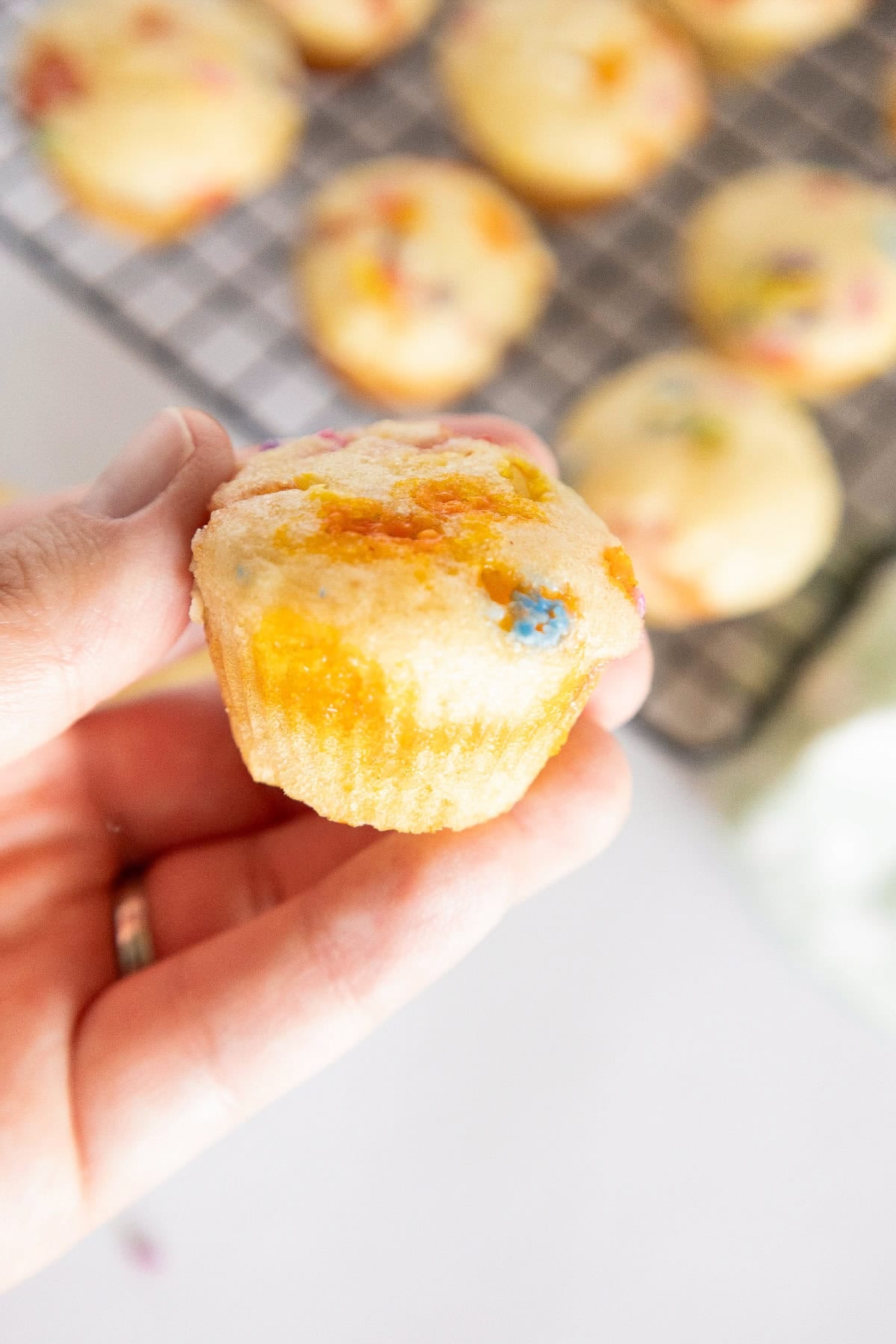 Hand holding a birthday cake mini muffin with colorful sprinkles and blurred muffins in the background.