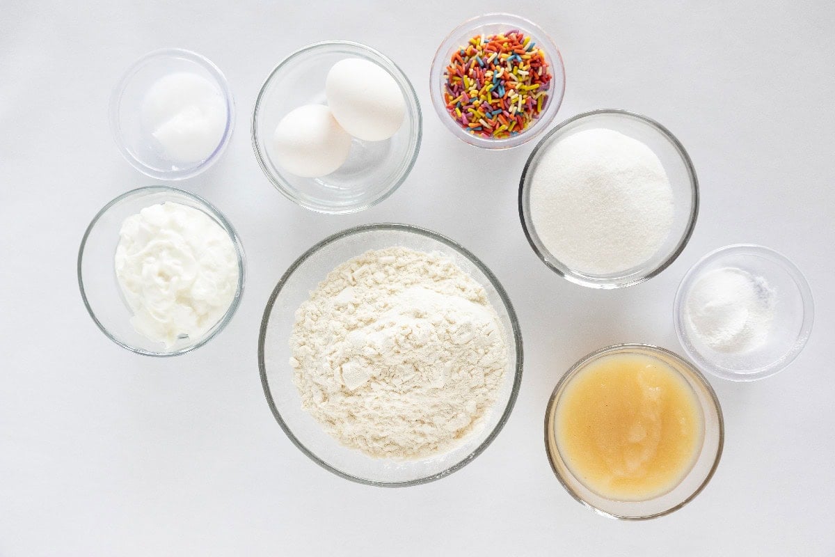 Ingredients for birthday cake mini muffins arranged in glass bowls, including flour, sugar, eggs, yogurt, melted butter, sprinkles, baking powder, and baking soda on a white background.