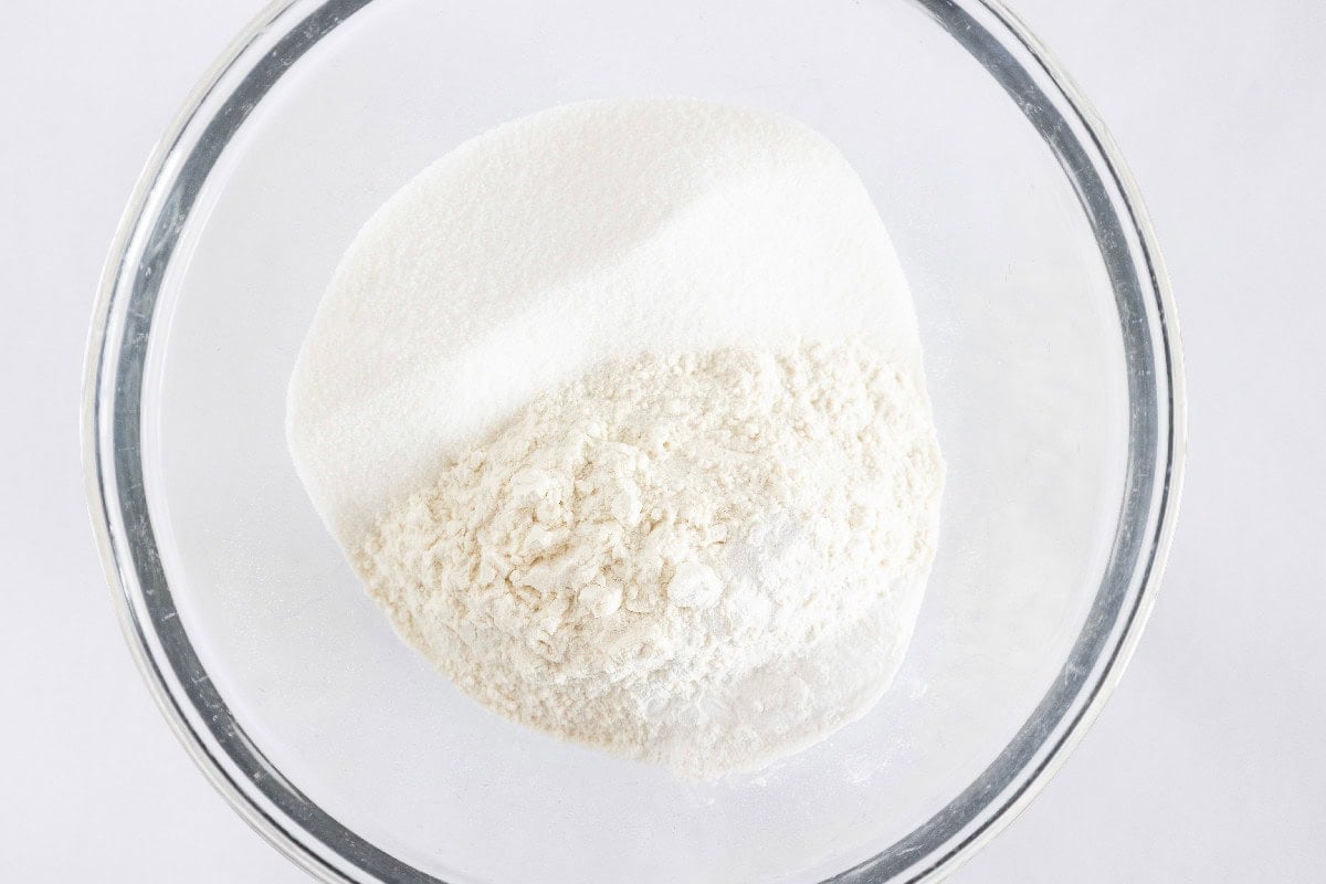 Dry ingredients for birthday cake mini muffins, including flour, sugar, baking powder, and baking soda mixed in a glass bowl on a white background.