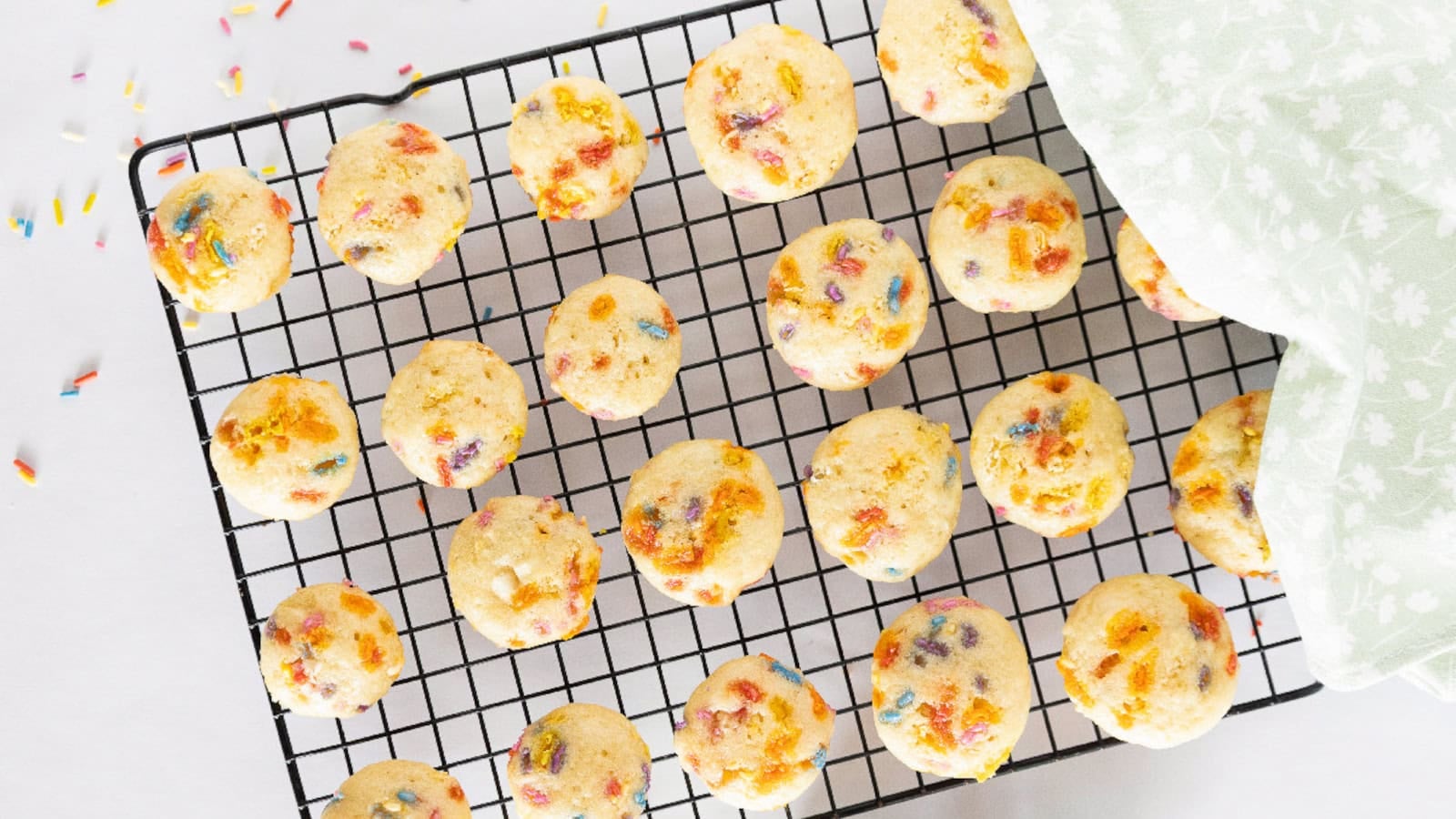 Birthday cake mini muffins are cooling on a wire rack with colorful rainbow sprinkles.