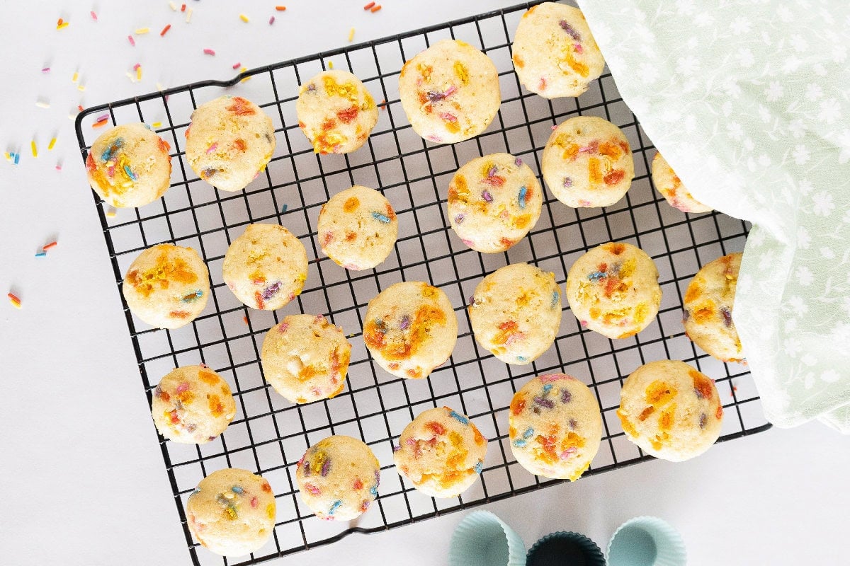 Birthday cake mini muffins are cooling on a wire rack with rainbow sprinkles visible on top.