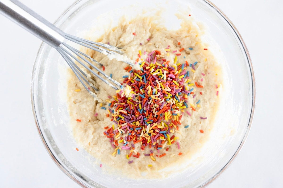 Rainbow sprinkles added to birthday cake mini muffin batter in a glass bowl.