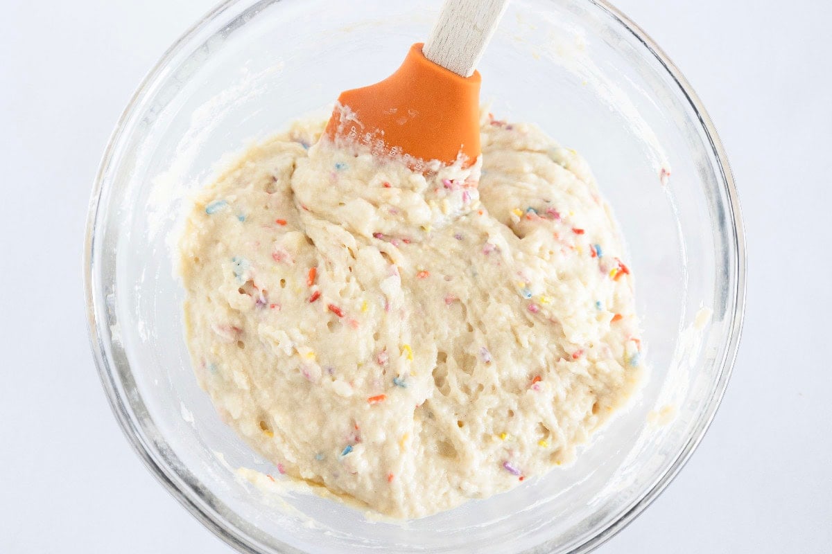 Folding rainbow sprinkles into birthday cake mini muffin batter with a spatula.