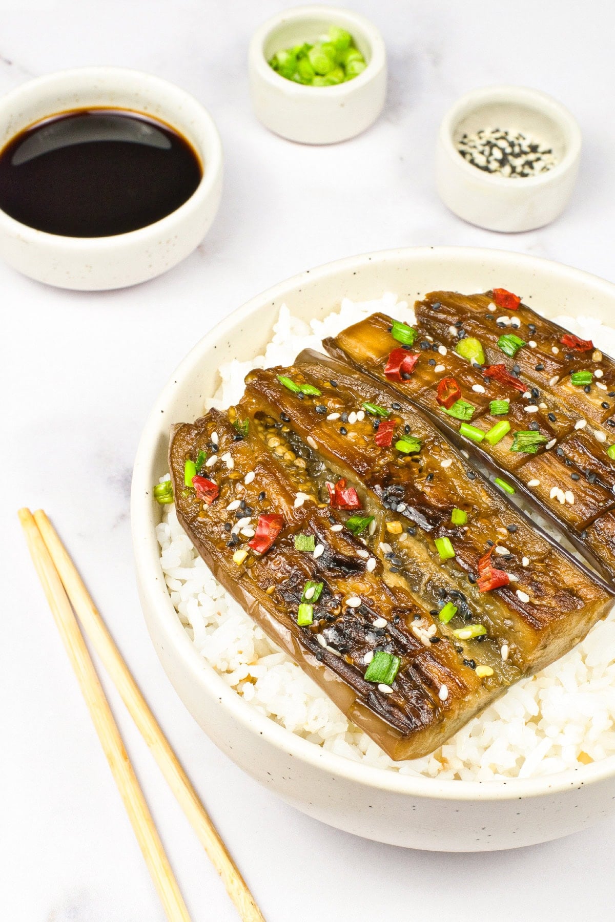 Side-angle shot of glazed vegan unagi eggplant served over steamed white rice with chopsticks and soy sauce on the side.