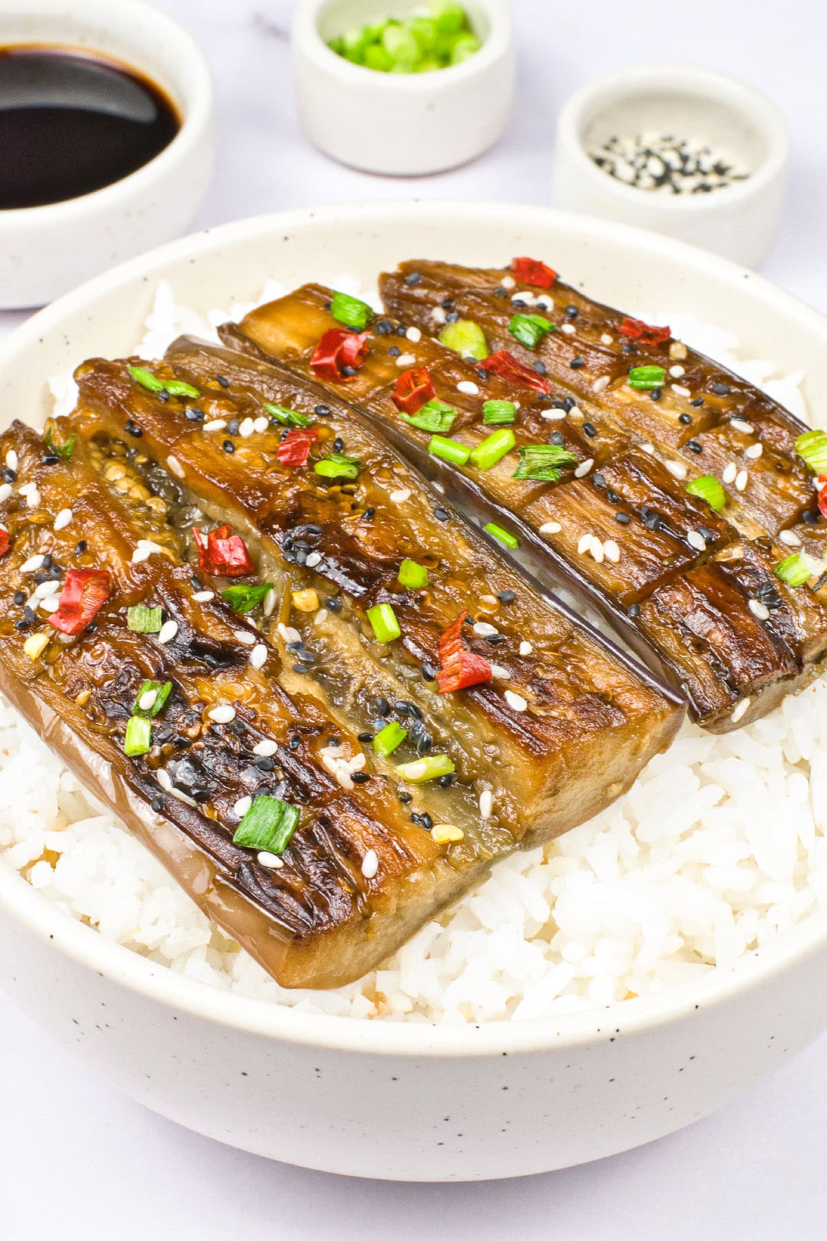 Close-up angled view of vegan unagi-style eggplant over white rice topped with sesame seeds, chopped green onions, and red chili pieces.
