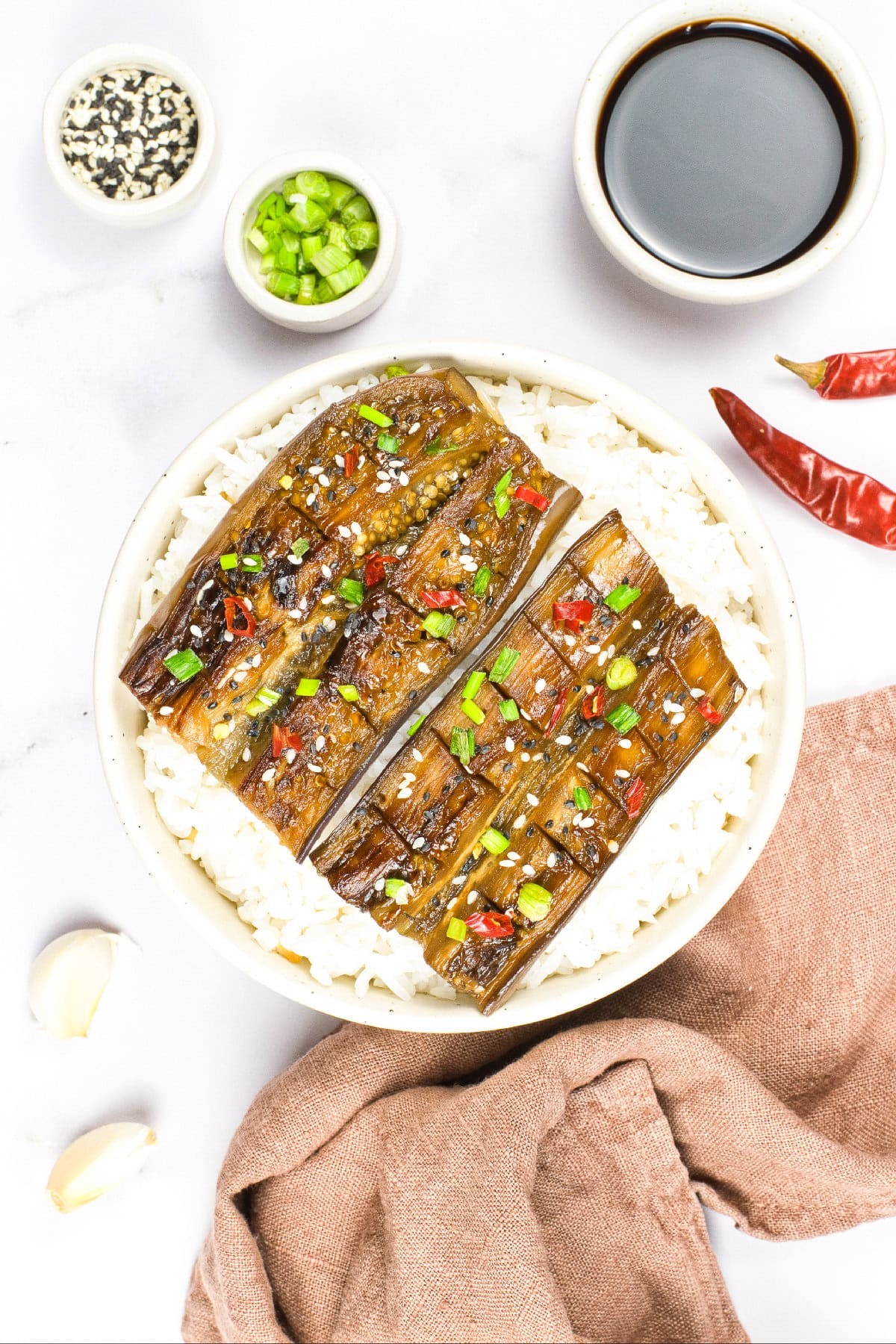 Overhead shot of vegan unagi-style eggplant served over white rice, garnished with sesame seeds, green onions, and red chili slices, with soy sauce and seasonings on the side.