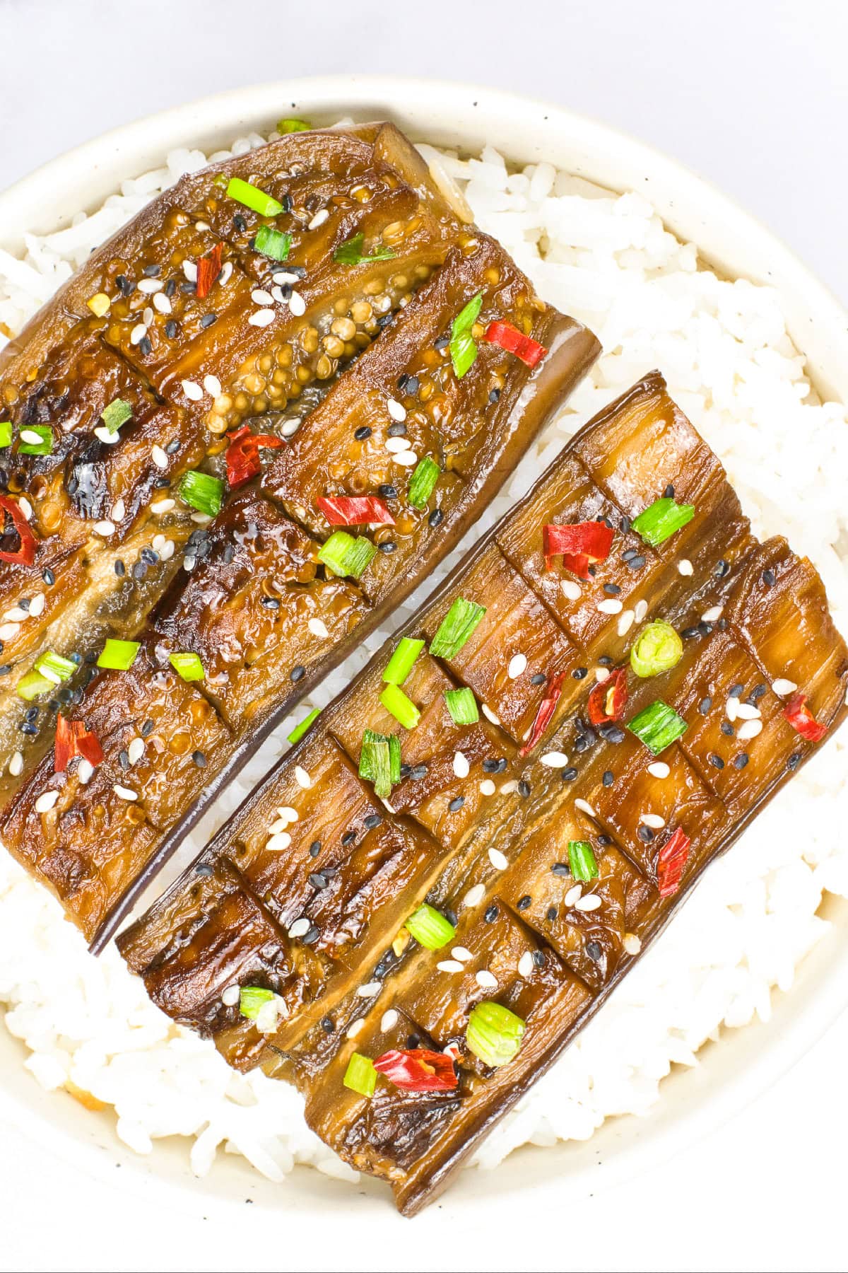 Close-up overhead view of glazed vegan unagi eggplant over white rice with sesame seeds, chopped green onions, and red chili garnish.