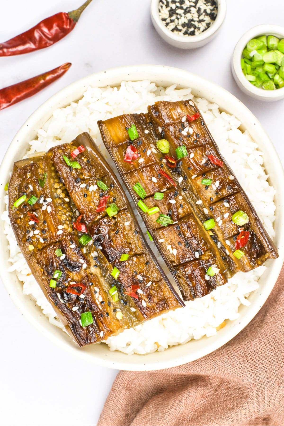 Close-up view of caramelized vegan unagi eggplant served over white rice with sesame seeds, chopped green onions, and red chili flakes.