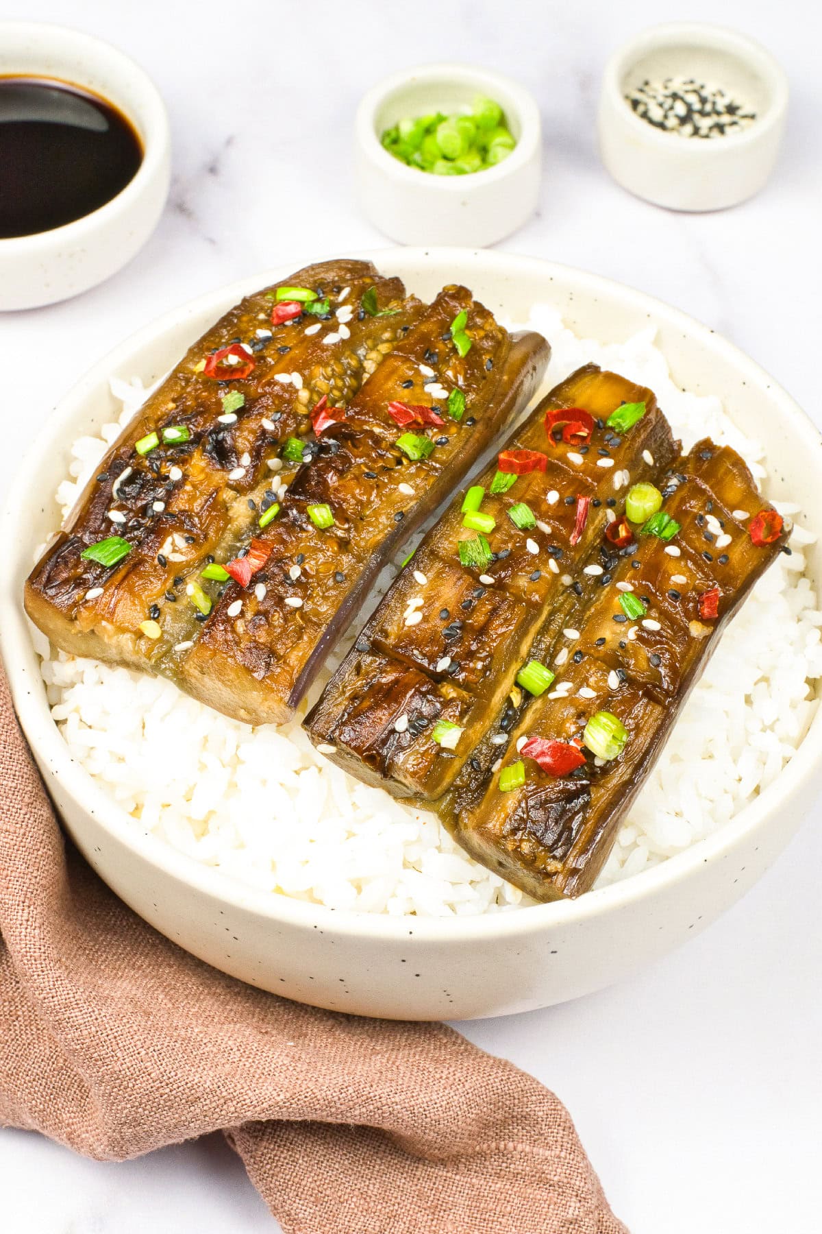 Angled view of vegan unagi eggplant over white rice with soy sauce bowls, sesame seeds, and green onion garnish in the background.