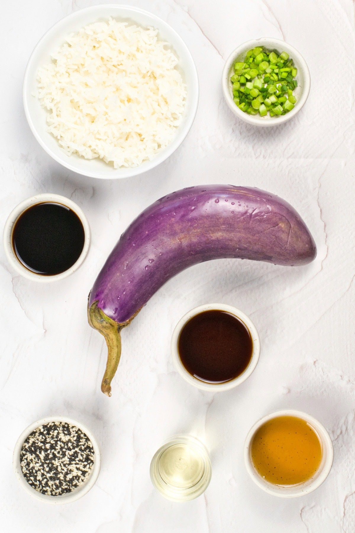 Ingredients for vegan unagi-style eggplant dish, including a whole purple eggplant, cooked white rice, chopped green onions, soy sauce, teriyaki-style sauce, sesame seeds, and cooking oil arranged in small bowls on a light marble surface.