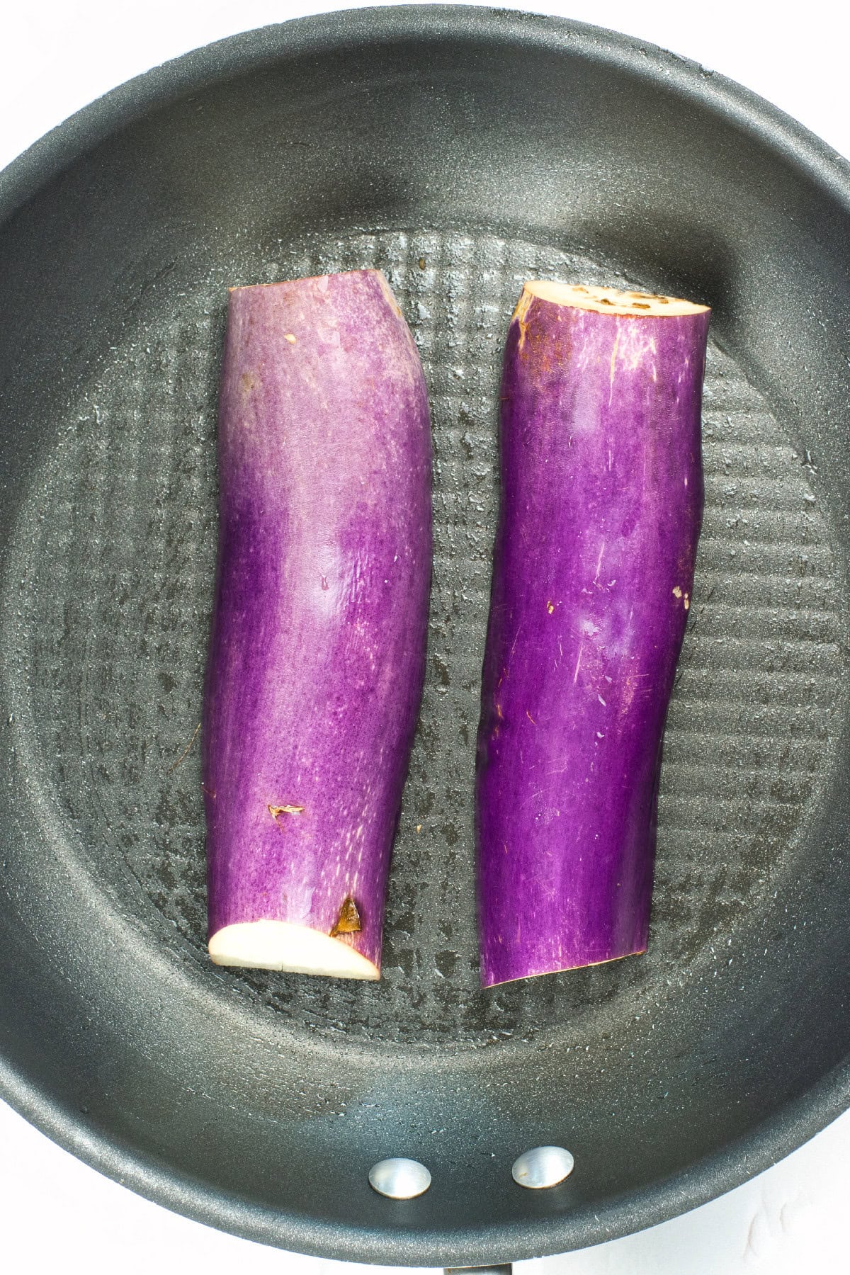 Two eggplant halves placed skin-side down in a lightly oiled skillet before cooking.