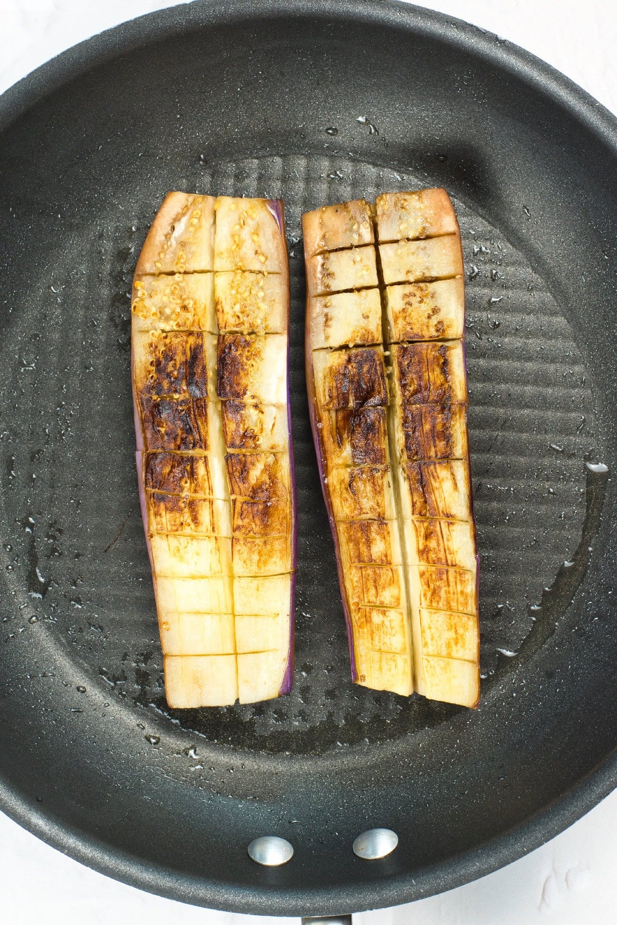 Scored eggplant halves cooking in a skillet with golden brown seared marks forming on the surface.