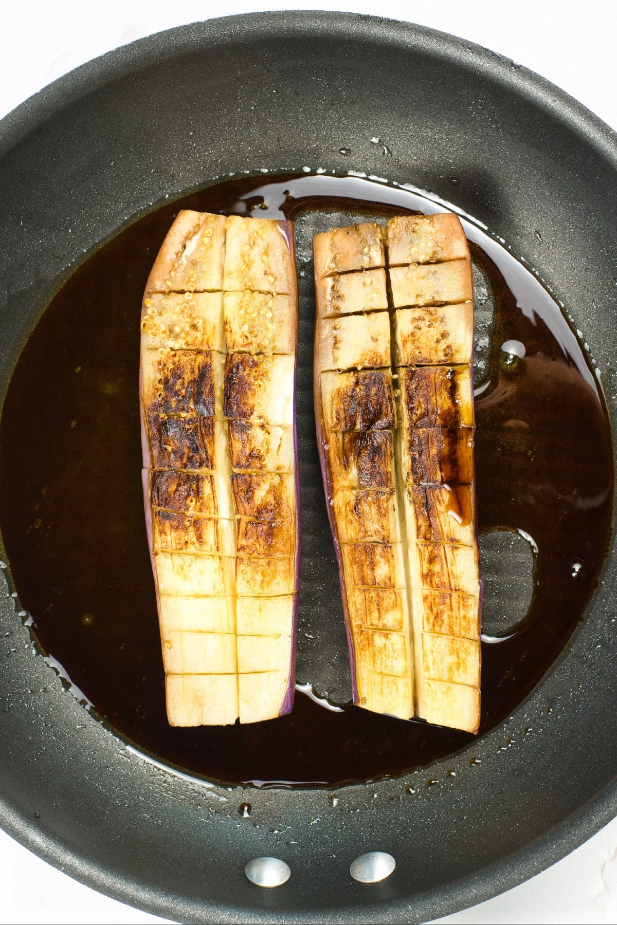 Cooked eggplant halves simmering in a savory vegan unagi sauce inside a skillet.