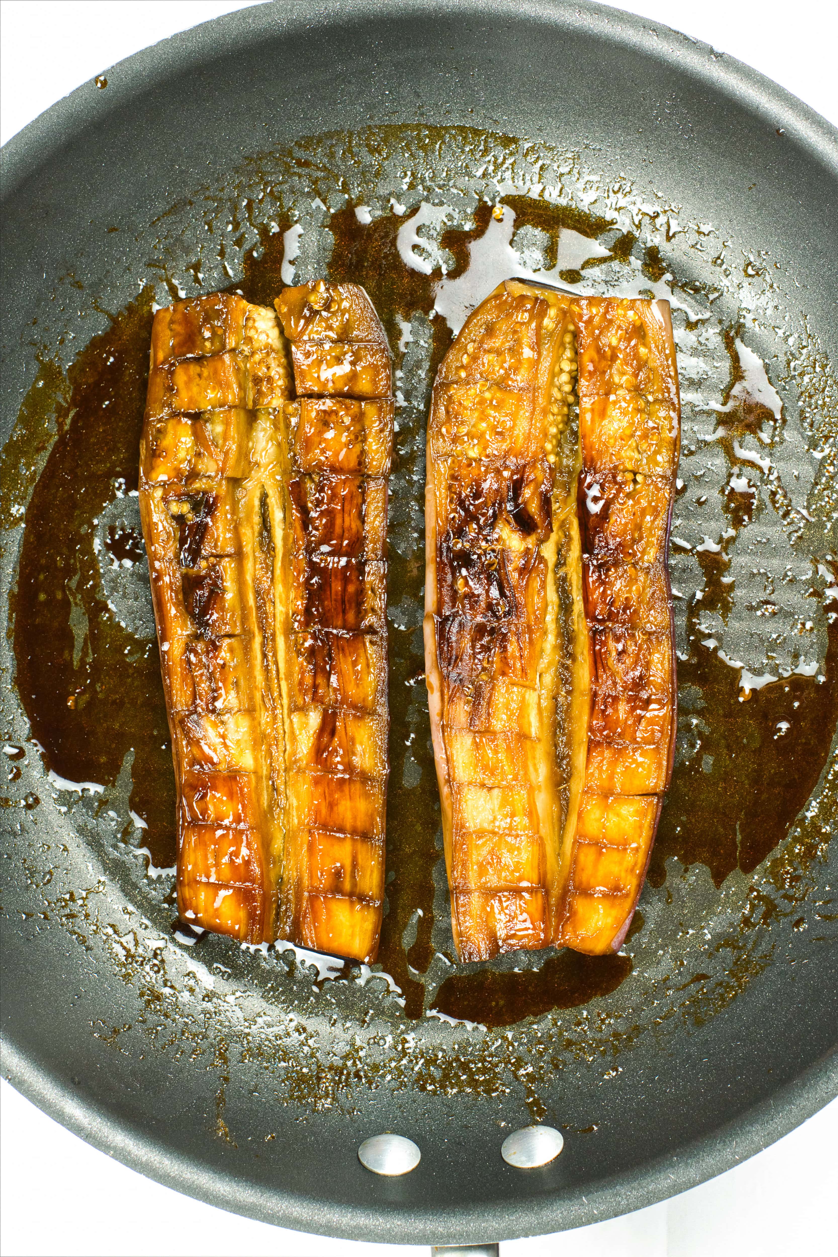 Glazed eggplant halves caramelizing in a skillet with thickened vegan unagi sauce.