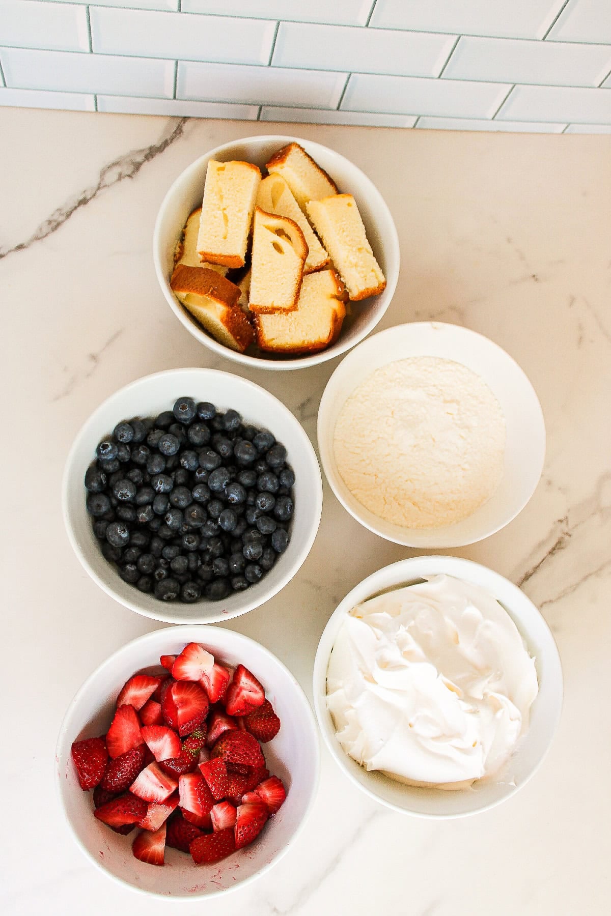 Ingredients for parfait cups including sliced strawberries, blueberries, vanilla pudding, whipped topping, and pound cake arranged in white bowls on a marble countertop.