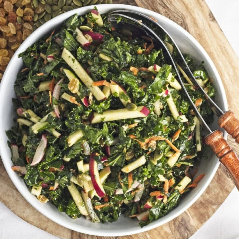 Kale apple salad with chopped kale, crisp red apple slices, shredded carrots, pumpkin seeds, and raisins tossed in a lemon miso dressing and served in a white bowl.