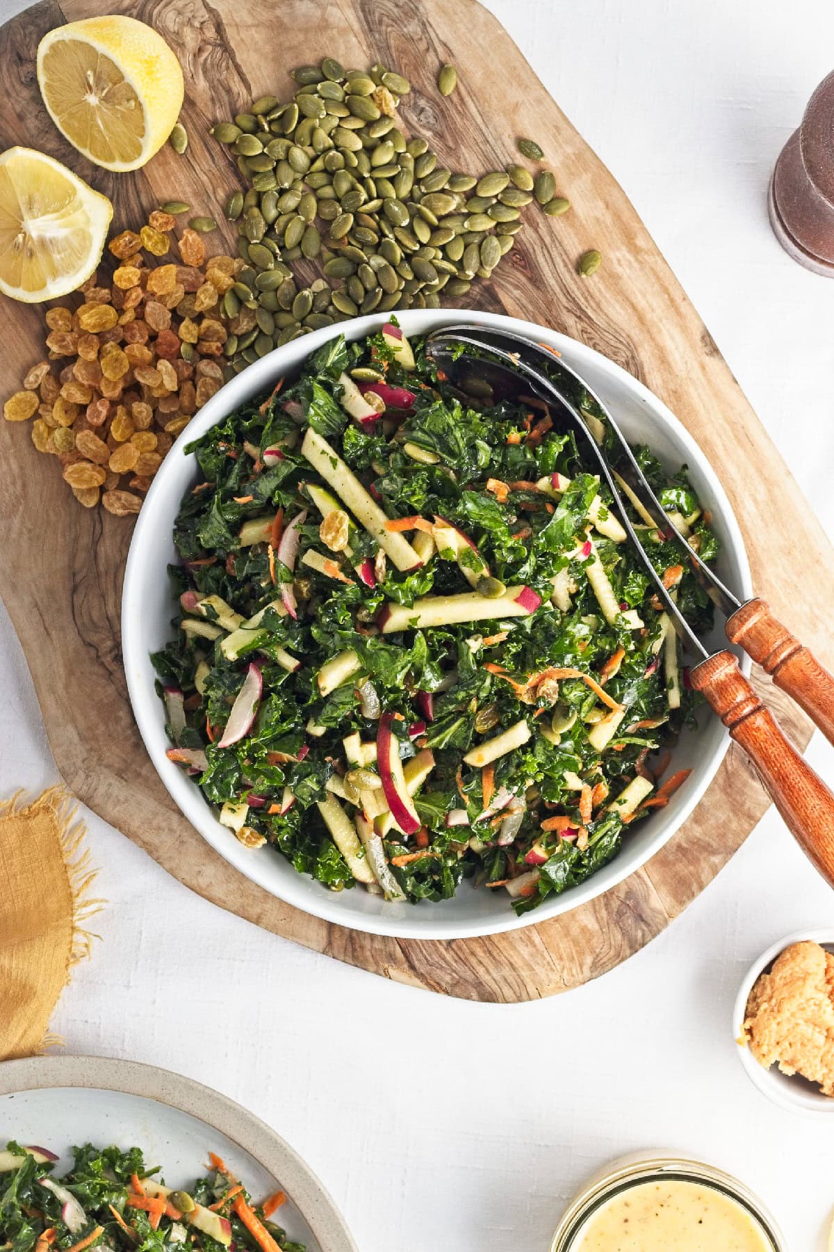 Kale apple salad with apples, carrots, radishes, pumpkin seeds, and raisins tossed in lemon miso dressing and served in a large white bowl.