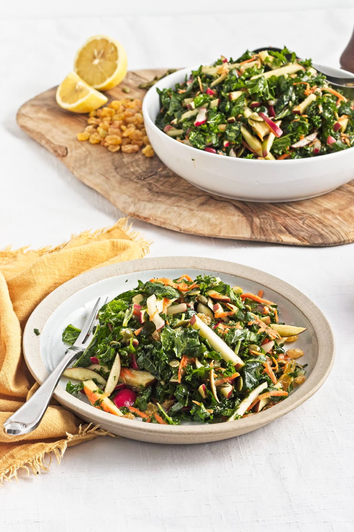 Served kale apple salad on a plate with fresh apples, carrots, radishes, pumpkin seeds, and raisins.