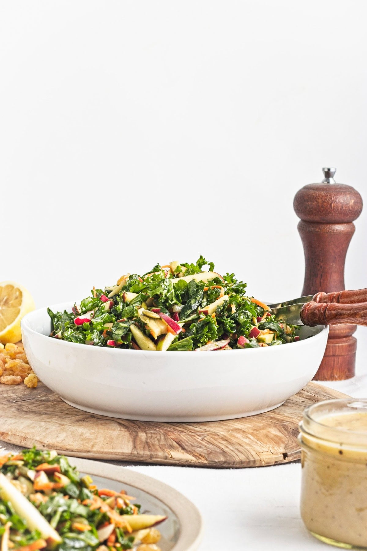 Side view of kale apple salad in a white serving bowl with homemade lemon miso dressing nearby.