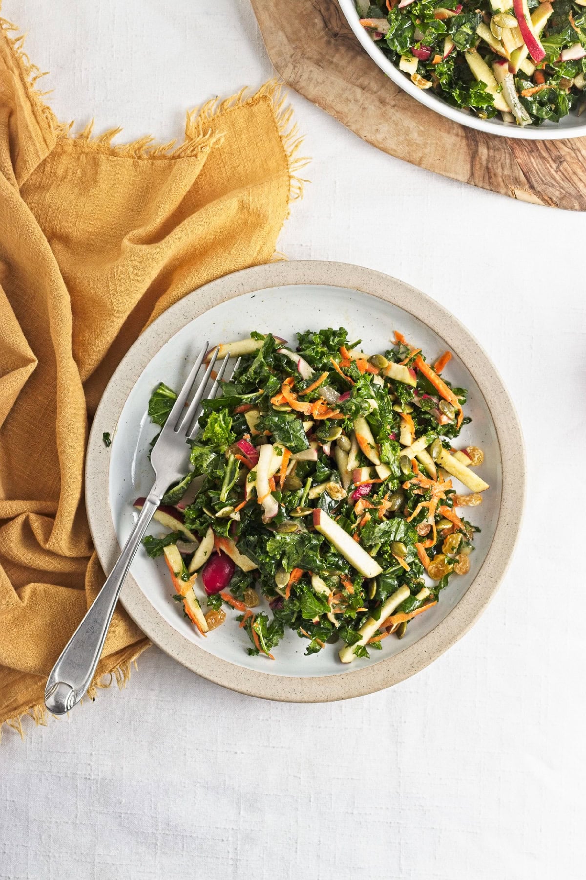 Single serving of kale apple salad on a plate with apples, carrots, radishes, pumpkin seeds, and raisins.