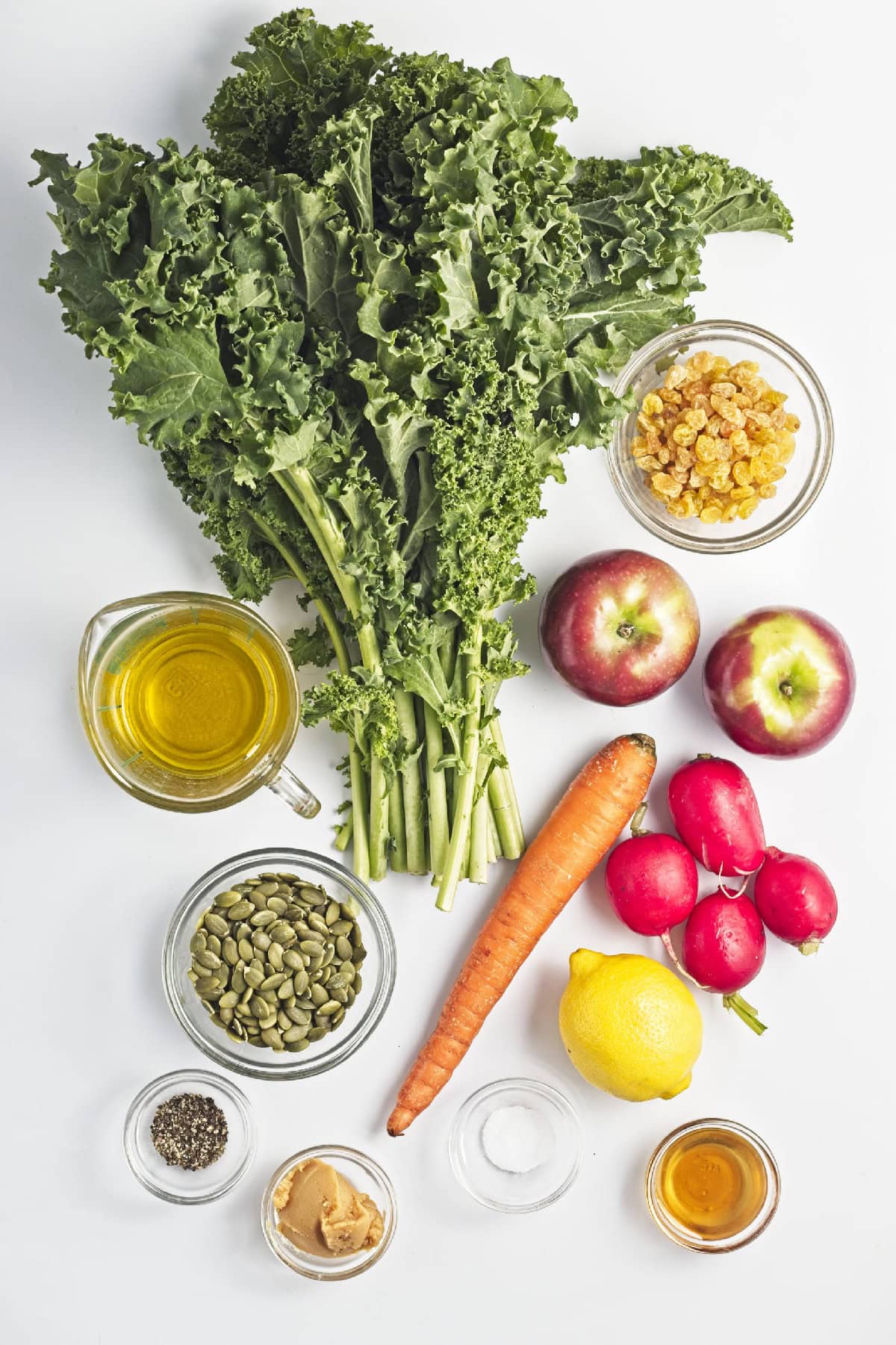 ngredients for kale apple salad including fresh kale, apples, carrot, radishes, pumpkin seeds, raisins, lemon, olive oil, miso paste, maple syrup, salt, and black pepper.