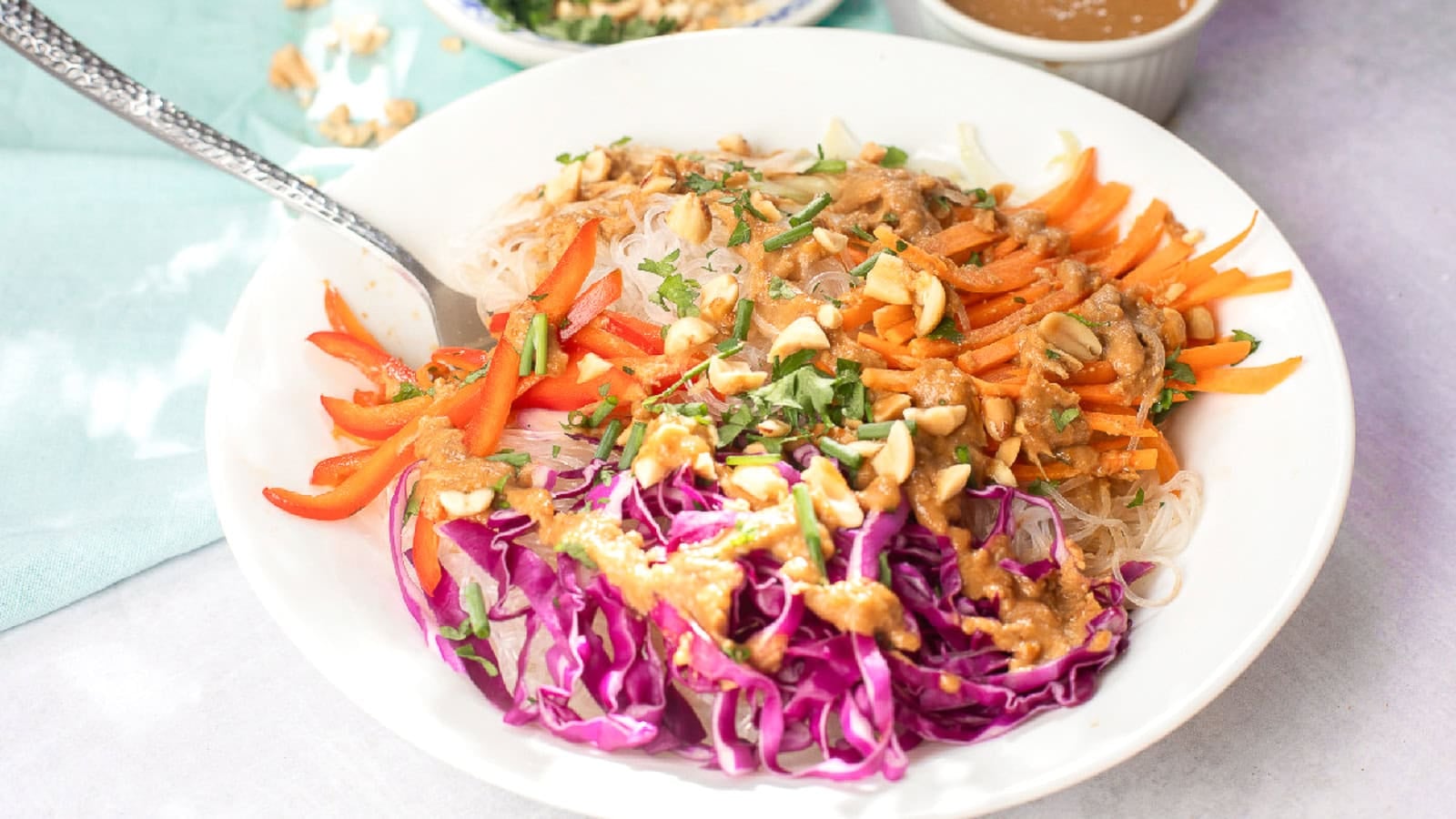 Spring roll in a bowl topped with rice noodles, shredded red cabbage, carrots, red bell peppers, chopped peanuts, fresh herbs, and creamy peanut sauce in a white bowl.