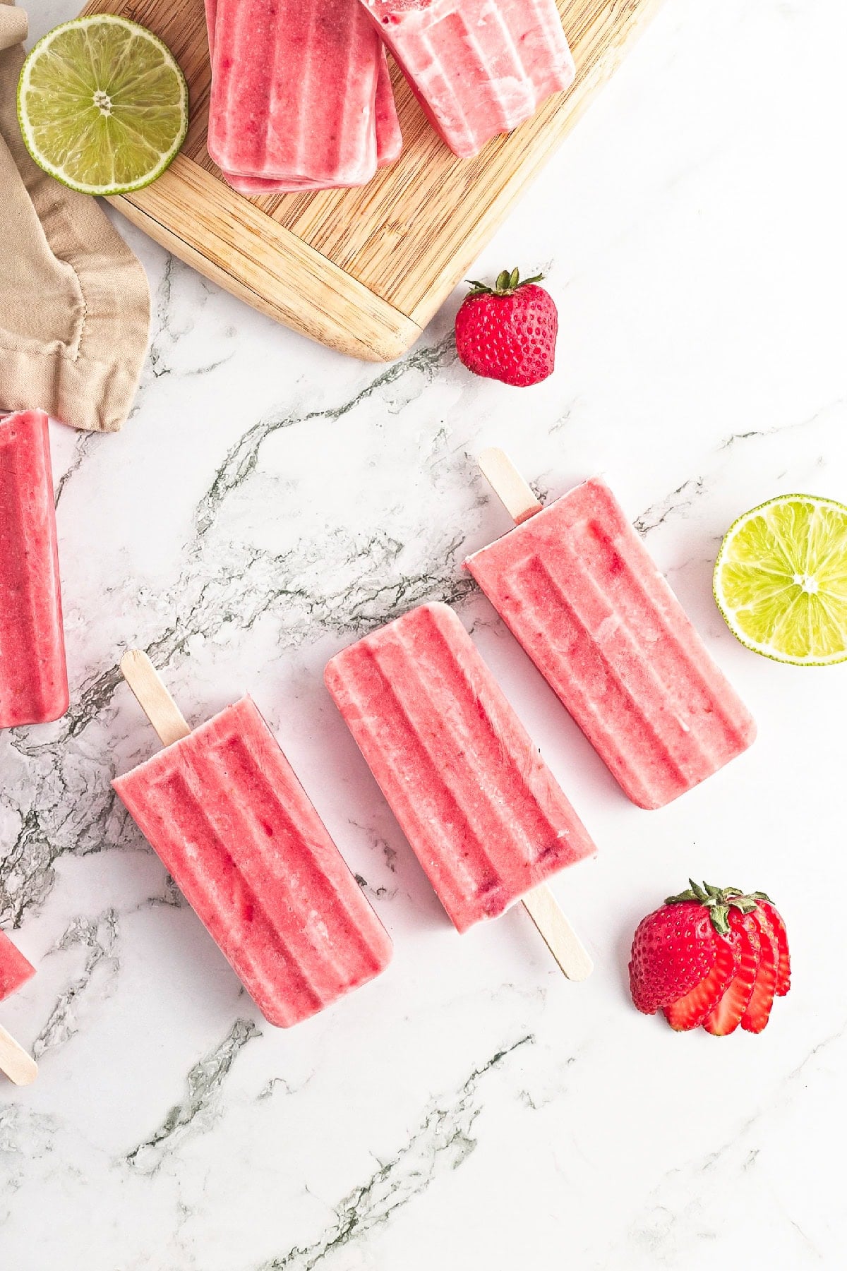 Fresh strawberry popsicles arranged on a marble countertop with strawberries and lime.
