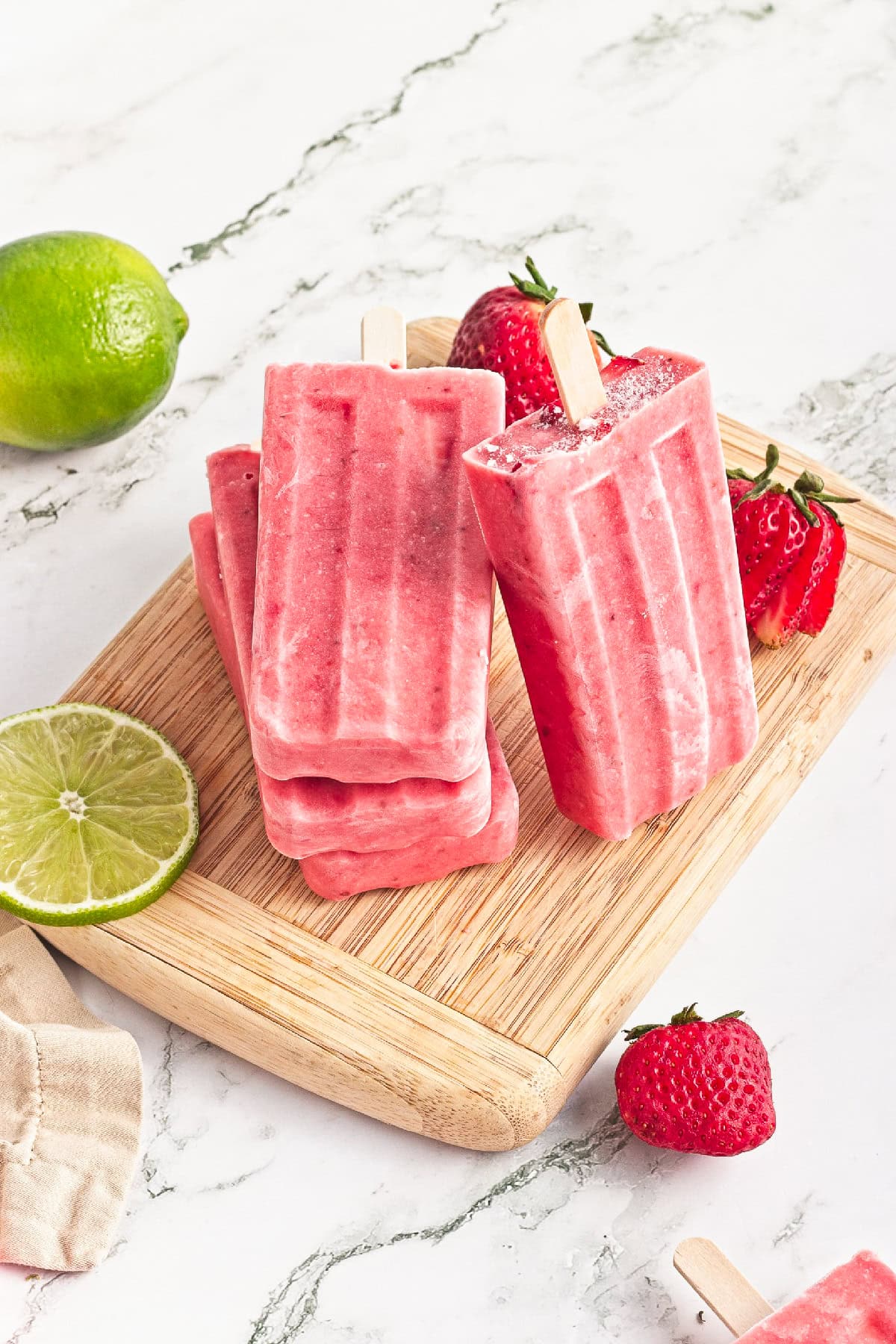 Creamy homemade strawberry popsicles stacked on a wooden board with fresh strawberries and lime.