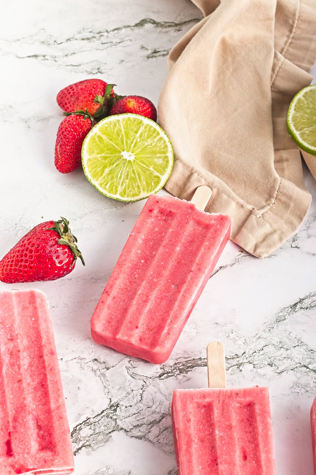 Homemade strawberry popsicle with fresh strawberries and sliced lime on a marble surface.