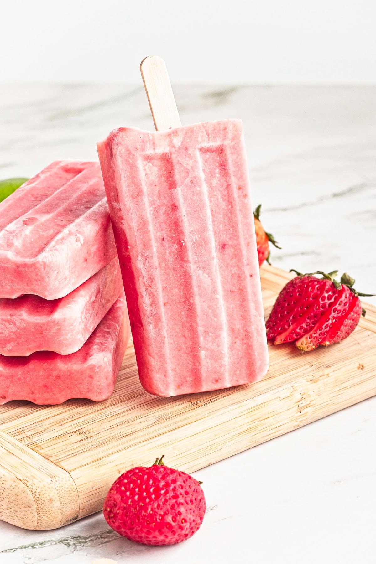 Single homemade strawberry popsicle on a wooden board with fresh strawberries and lime.